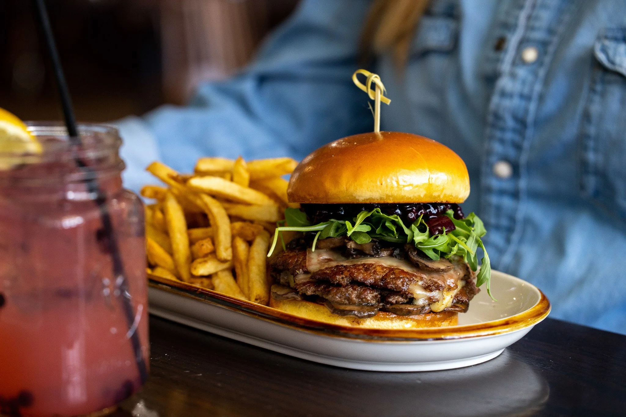 A gourmet burger with multiple beef patties, cheese, greens, a sauce, and berries, served with French fries on a white platter, with a drink in a mason jar nearby.