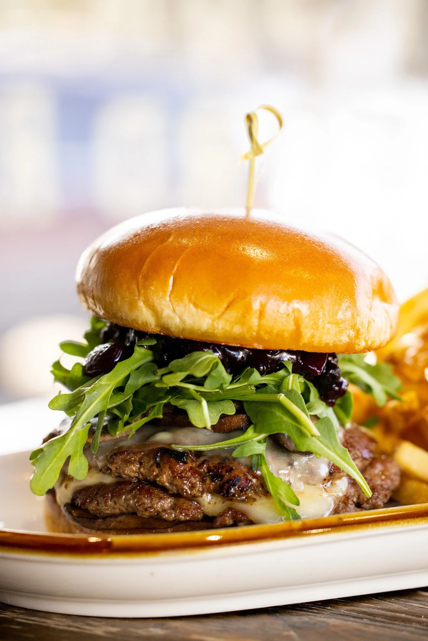 Close-up of a gourmet cheeseburger with arugula, black sauce, melted cheese, and a toasted bun, served with fries on a white plate.