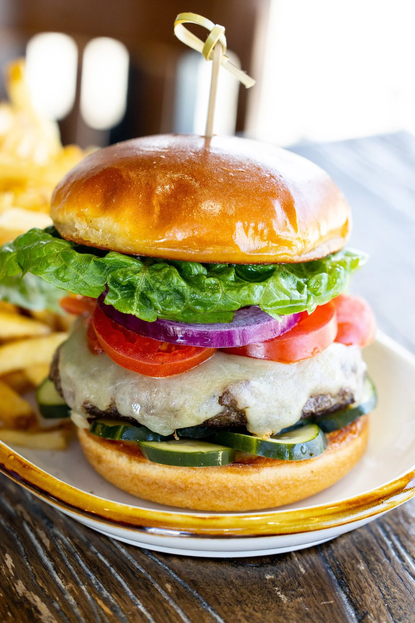 A close-up of a cheeseburger with lettuce, tomato, onion, cucumber slices, melted cheese, and a brioche bun, served on a plate with some fries in the background.