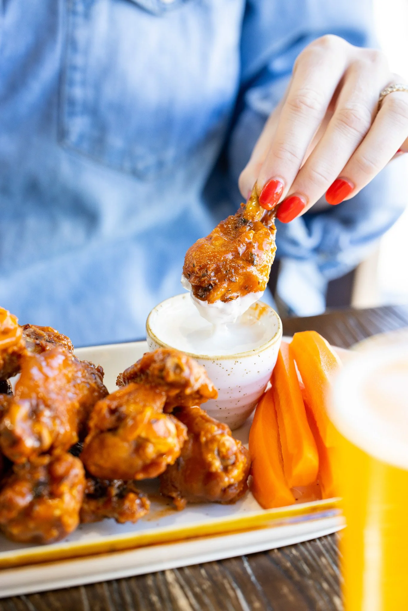 Person dipping a chicken wing into ranch dressing with more chicken wings and raw carrots on a plate.