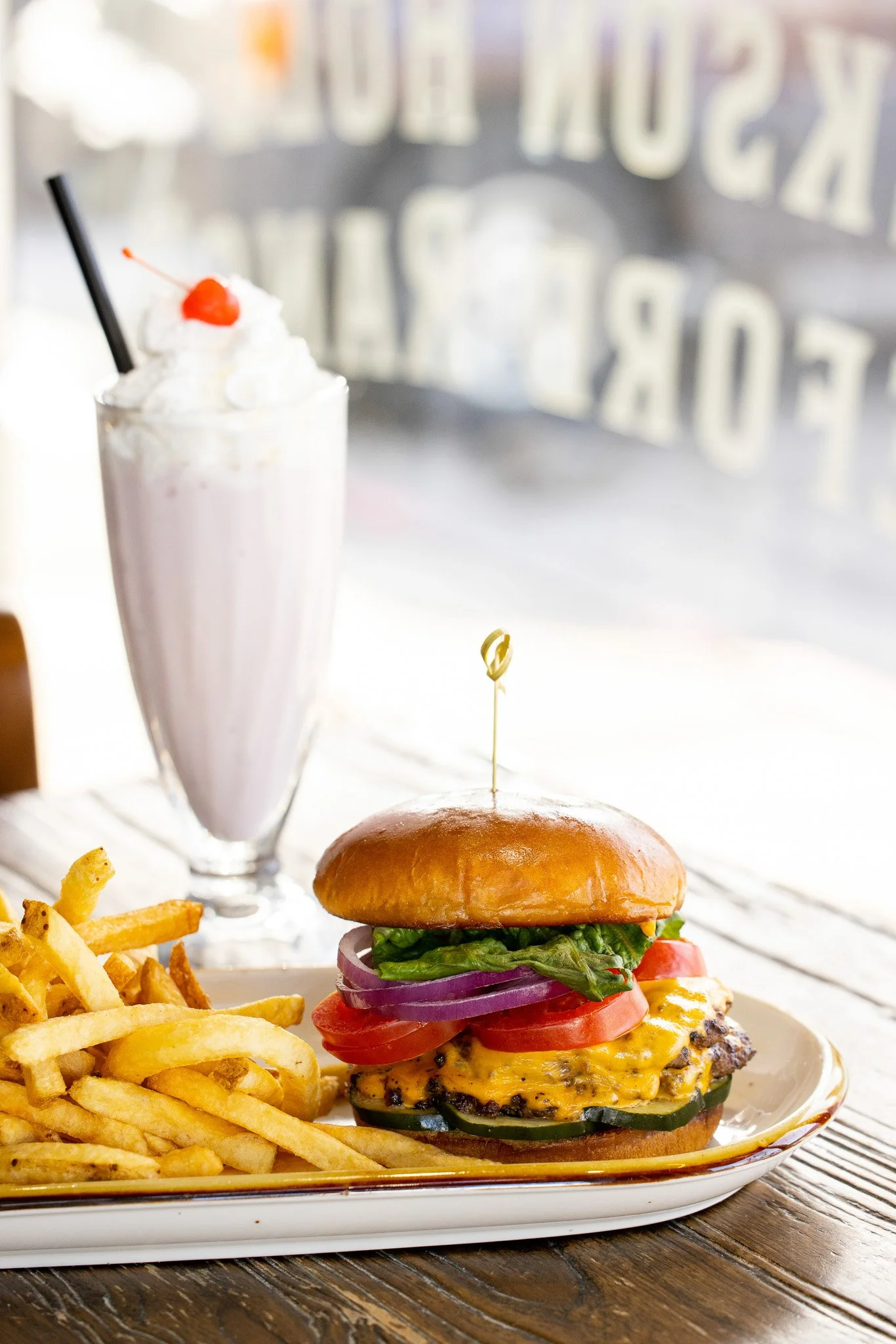 A cheeseburger with lettuce, tomato, onion, pickles, and cheese on a bun, served with French fries and a milkshake with whipped cream and a cherry, on a white plate in a restaurant setting.