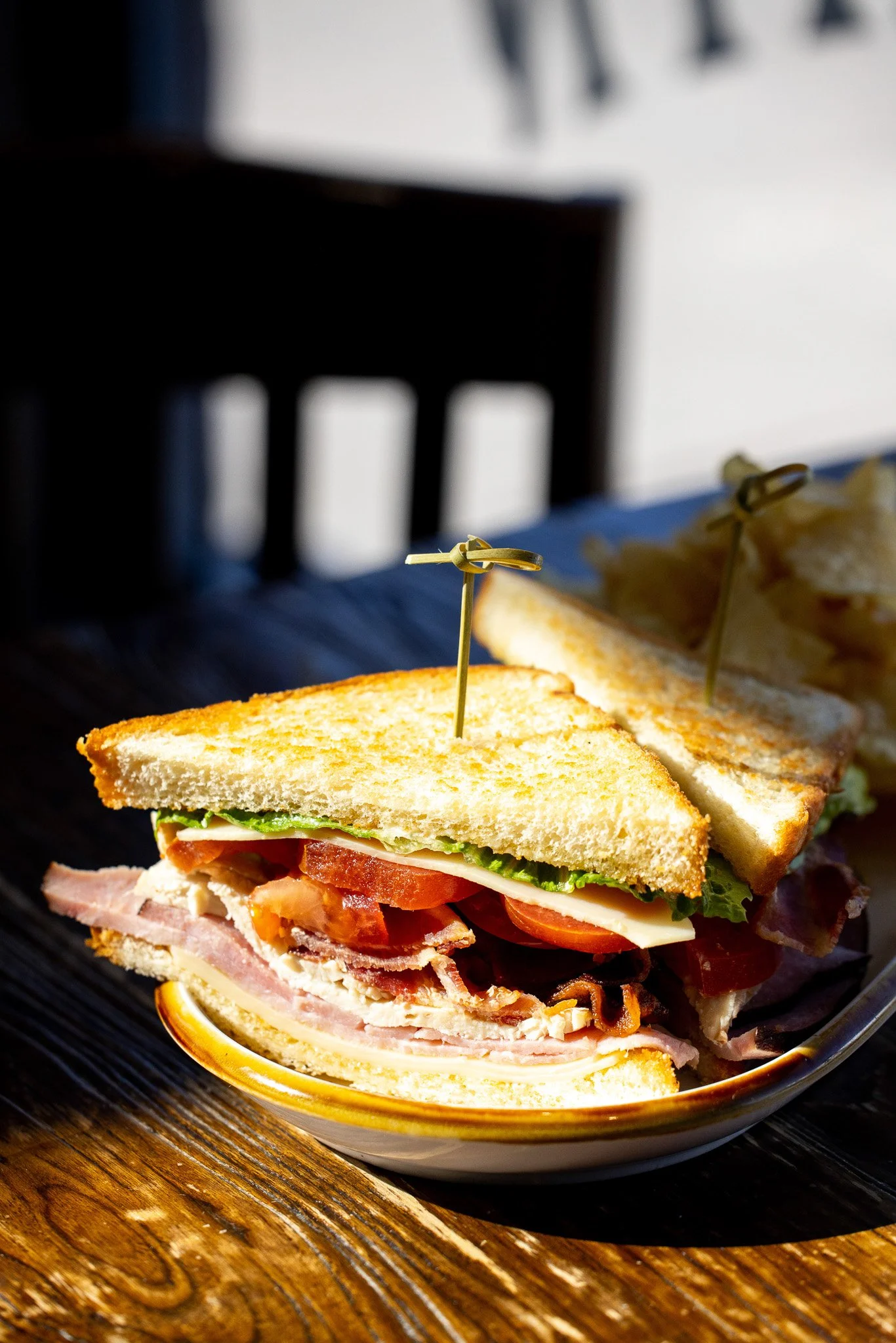 Club sandwich with bacon, lettuce, tomato, and turkey on toasted bread, skewered with a pick, served with potato chips on a patterned plate.