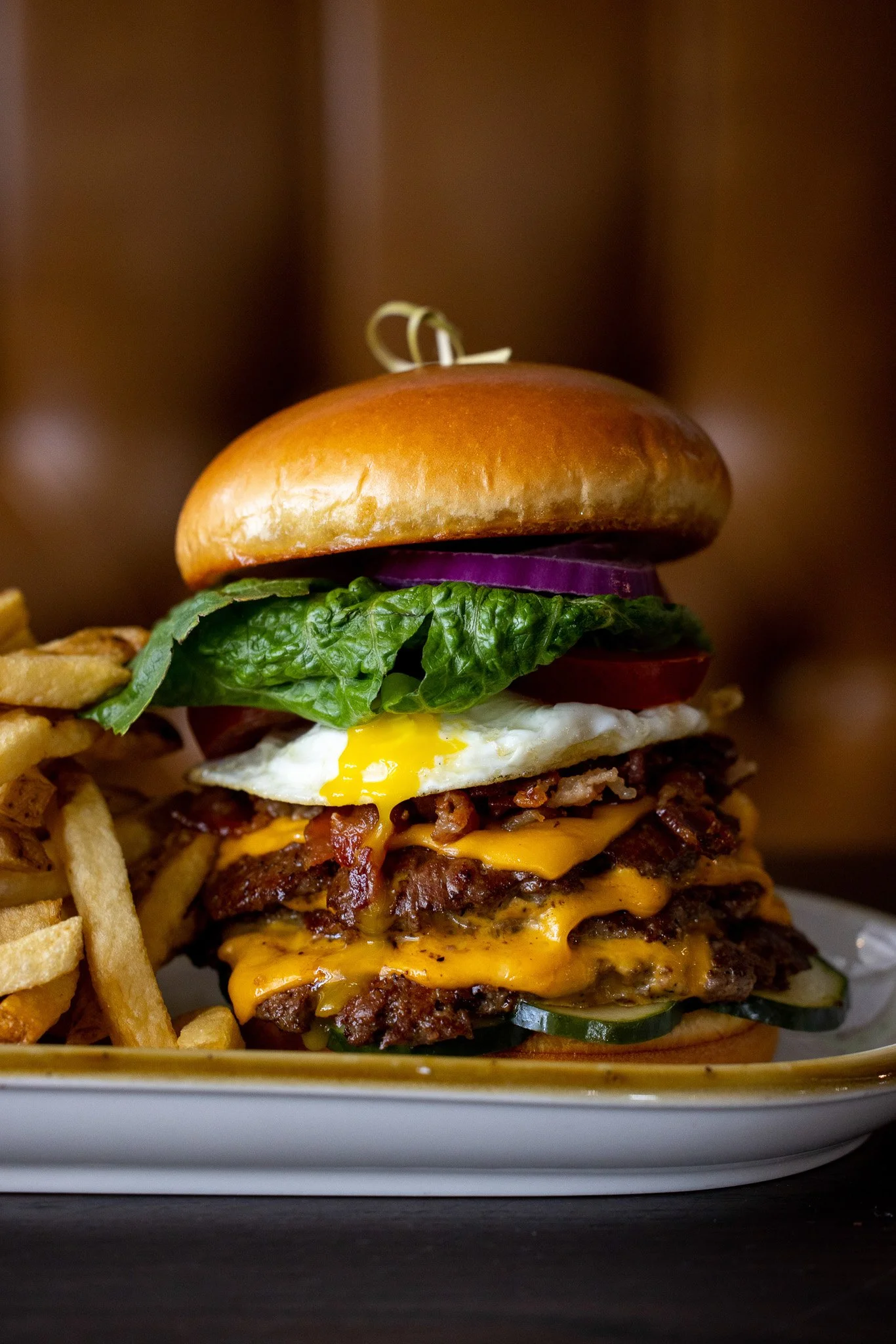 Close-up of a towering cheeseburger with lettuce, tomato, red onion, fried egg, two beef patties with melted cheddar cheese, and a toasted bun, served with a side of French fries.