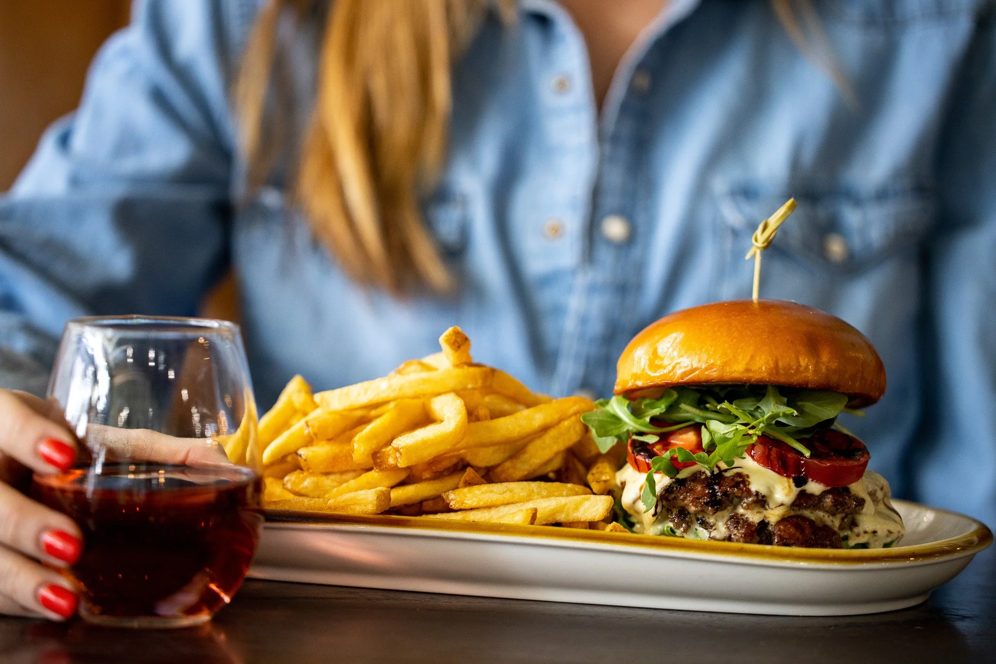 A person with red nail polish holding a glass of red wine, sitting in front of a plate with French fries and a cheeseburger topped with lettuce, tomato, and other toppings.