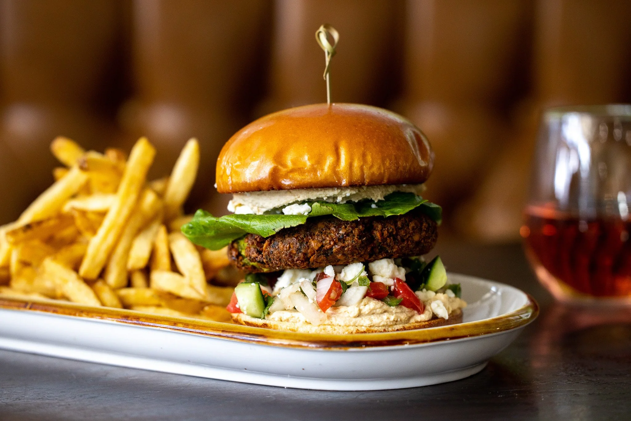 A plate of food featuring a large burger with lettuce, a fried patty, tomato, cucumber, cheese, and a bun, served with French fries and a glass of red beverage in the background.
