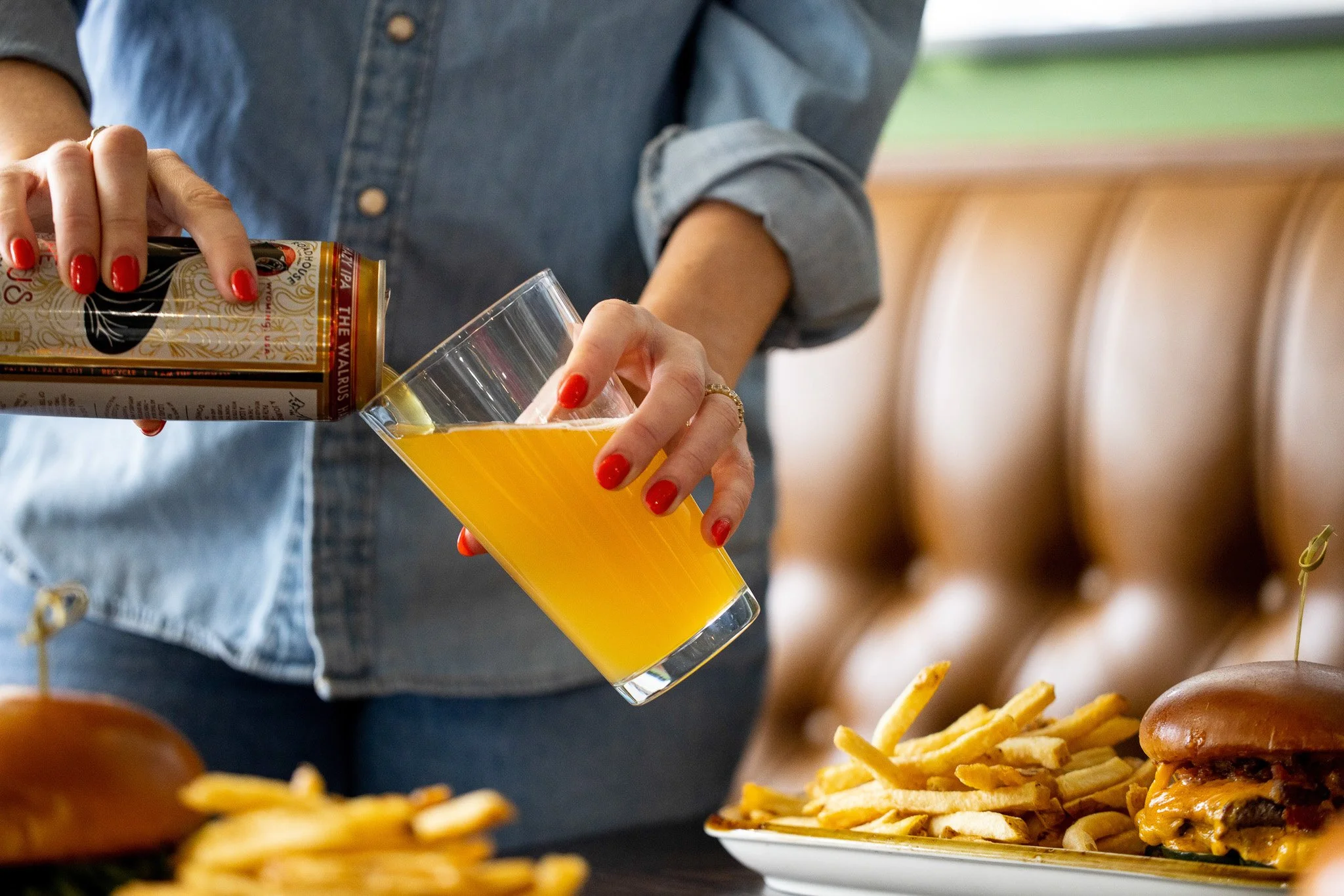 A person with red painted nails pouring a beer from a can into a glass. There is a plate of French fries and a cheeseburger with French fries on the table in front of them.