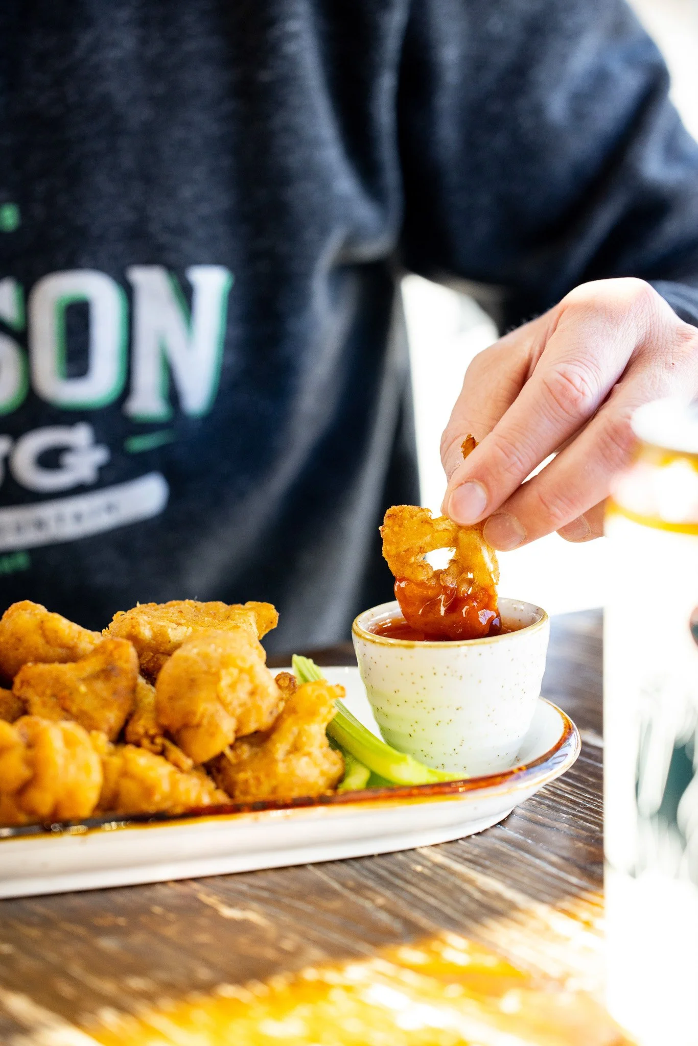 Person dipping a piece of fried chicken into hot sauce with a plate of fried chicken and celery stalks nearby.
