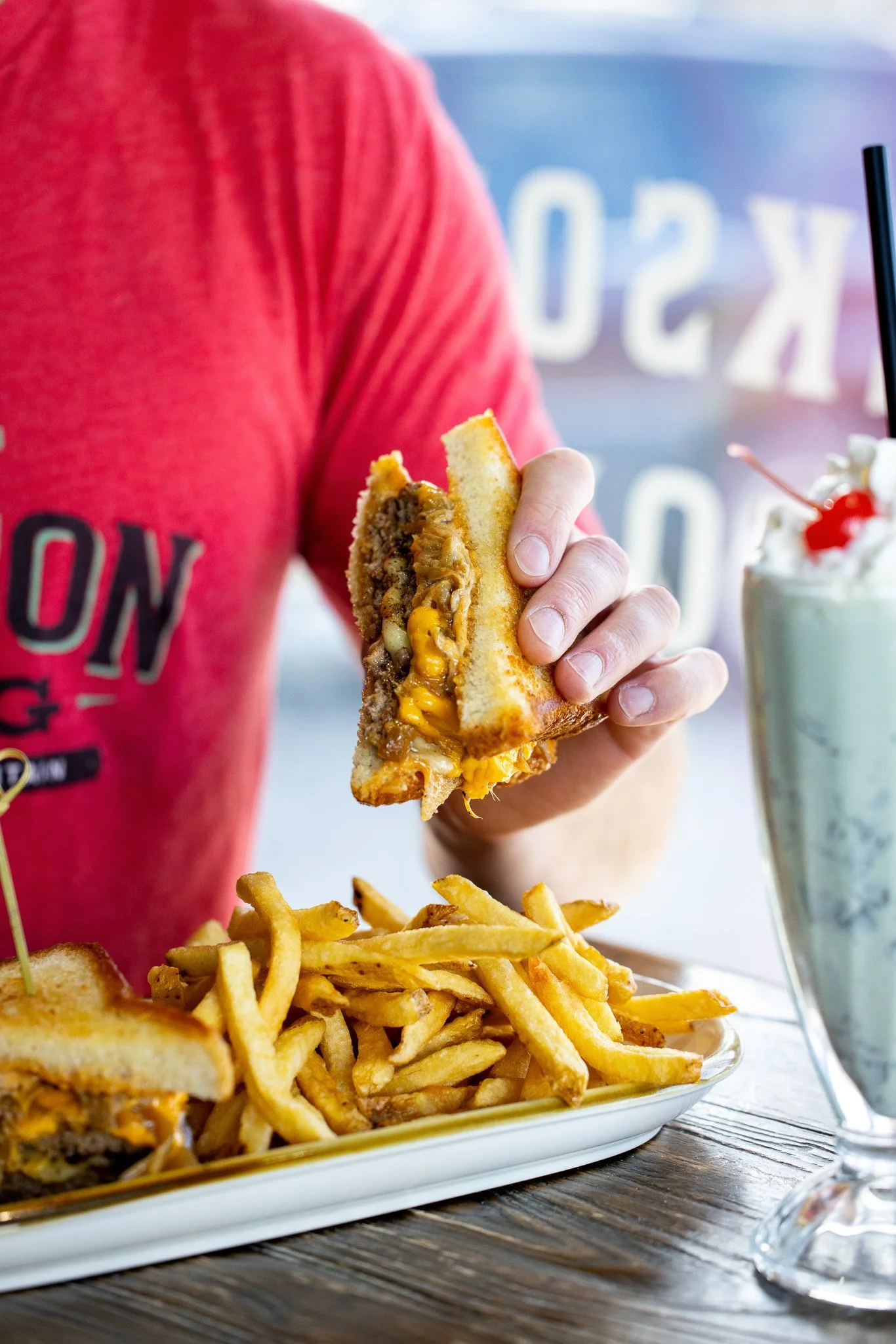 Person holding a cheeseburger with fries on a plate and a milkshake on a table.