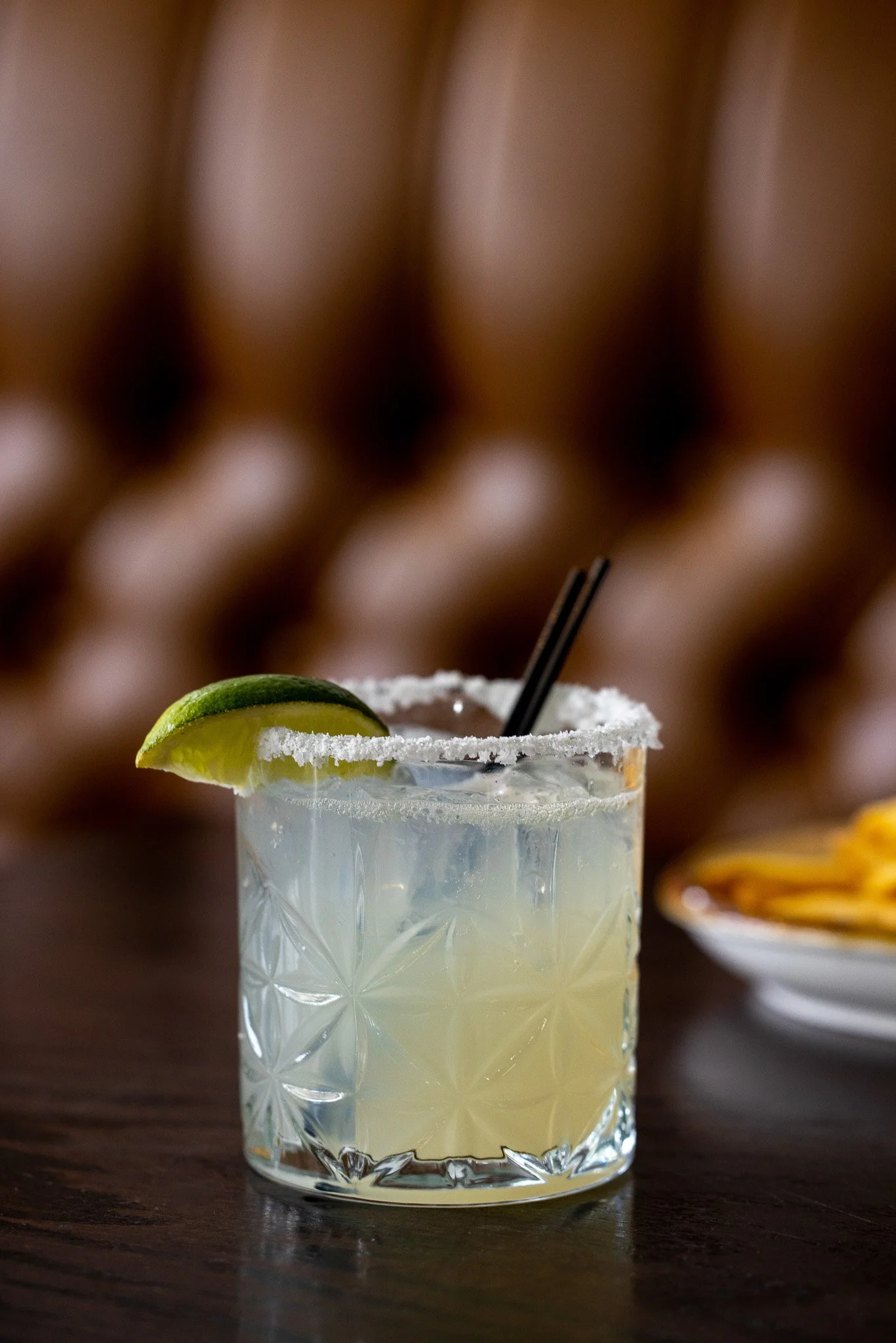 A glass of margarita with salt rim, lime wedge, and two black straws on a dark wooden surface, with a blurry background.