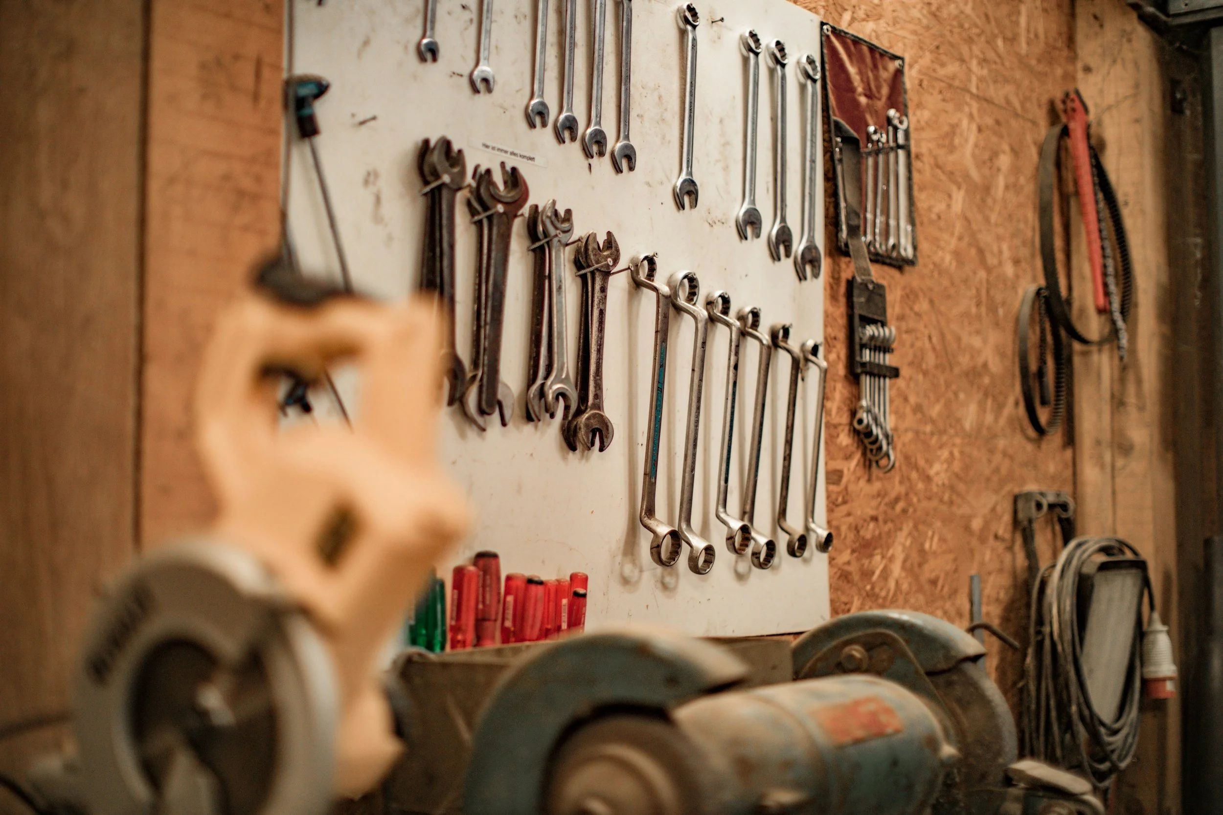 A workshop wall with various wrenches and tools hanging on a pegboard, along with power tools, containers, and cords on a workbench.