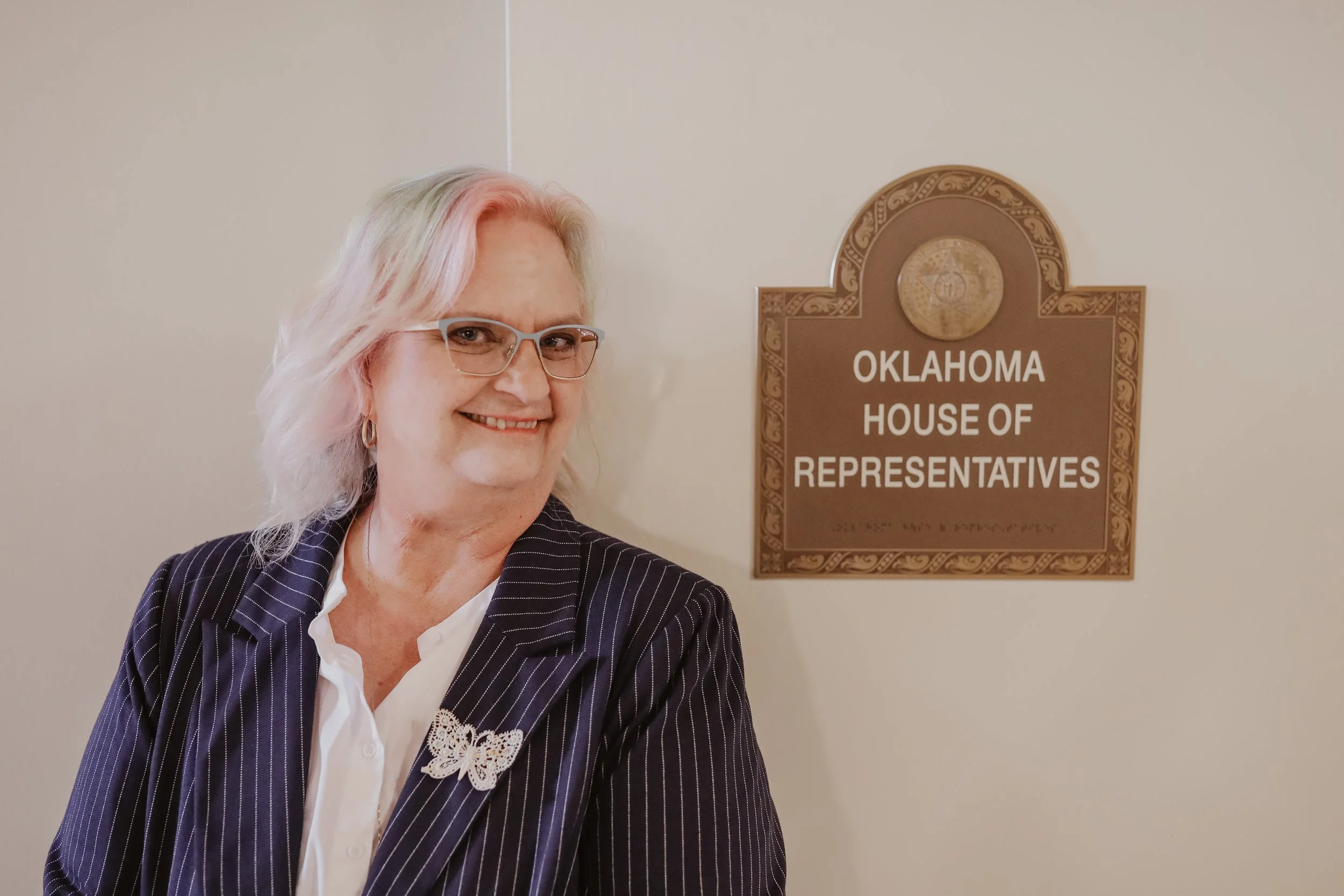 A smiling woman with pink hair, glasses, and gold earrings standing beside a sign for the Oklahoma House of Representatives.