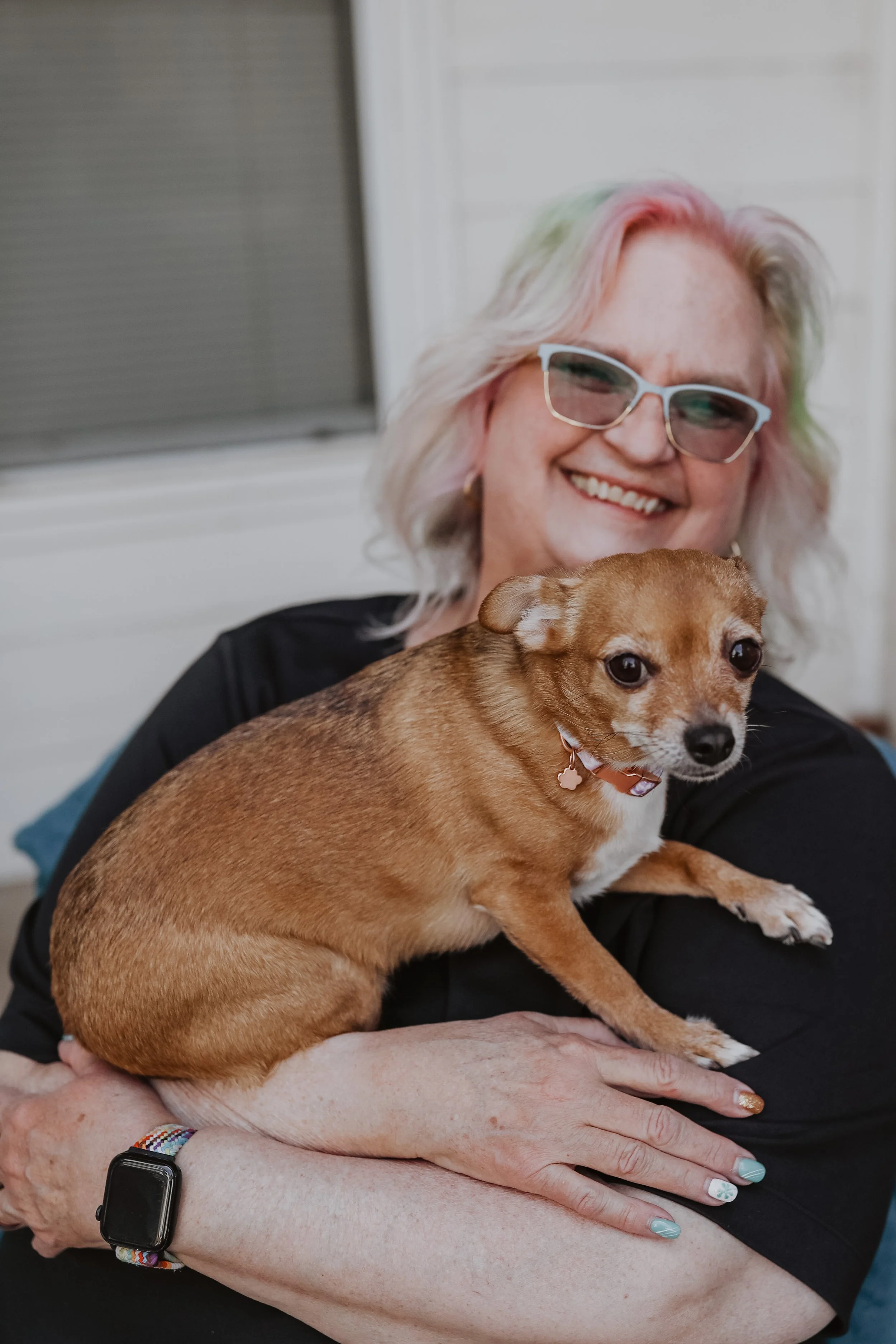 A woman,Vicki sits, smiling and holding a small brown dog, Iris in her arms.