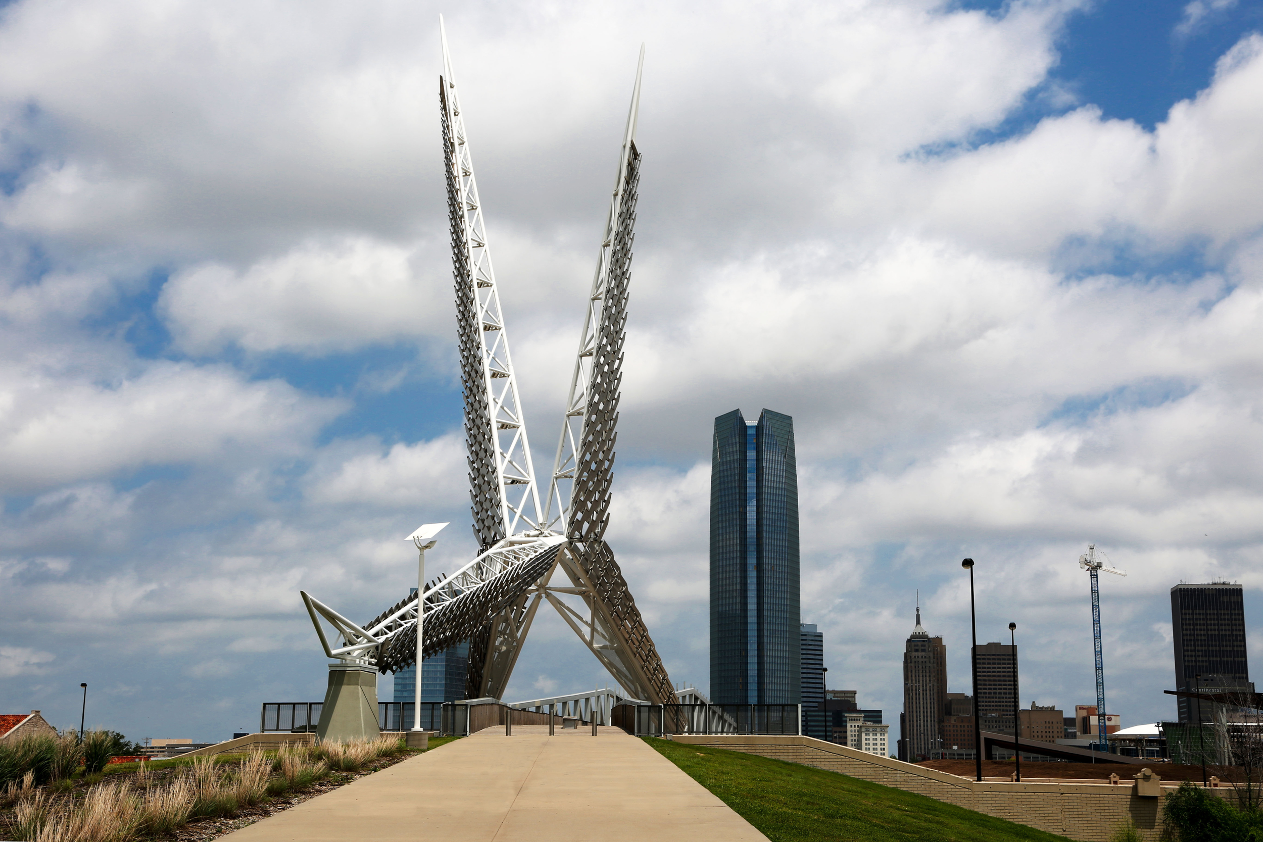 A modern sculpture resembling  wings in front of a city skyline with tall glass buildings and scattered clouds in the sky.