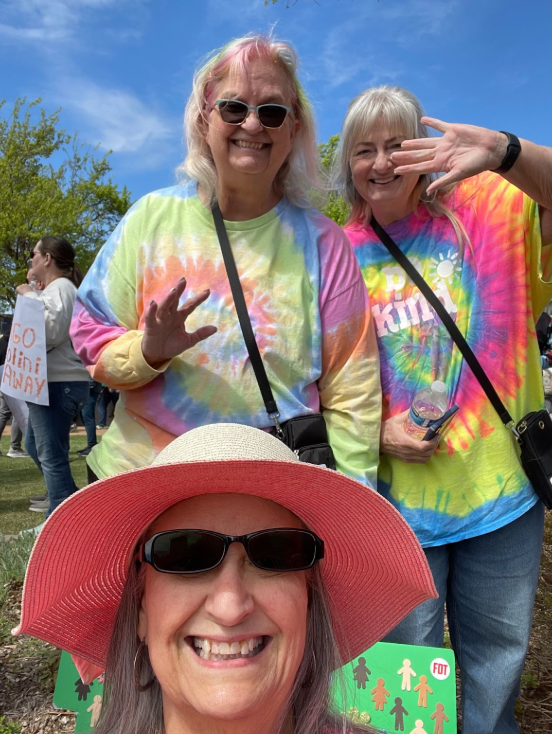 Three women smiling and waving at an outdoor event, two wearing colorful tie-dye shirts and one wearing a large pink floppy hat and sunglasses, with a crowd and trees in the background.