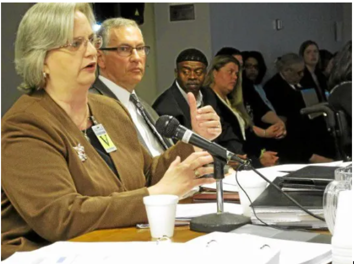 A woman speaking into a microphone at a panel discussion with several people seated behind her at a conference table.