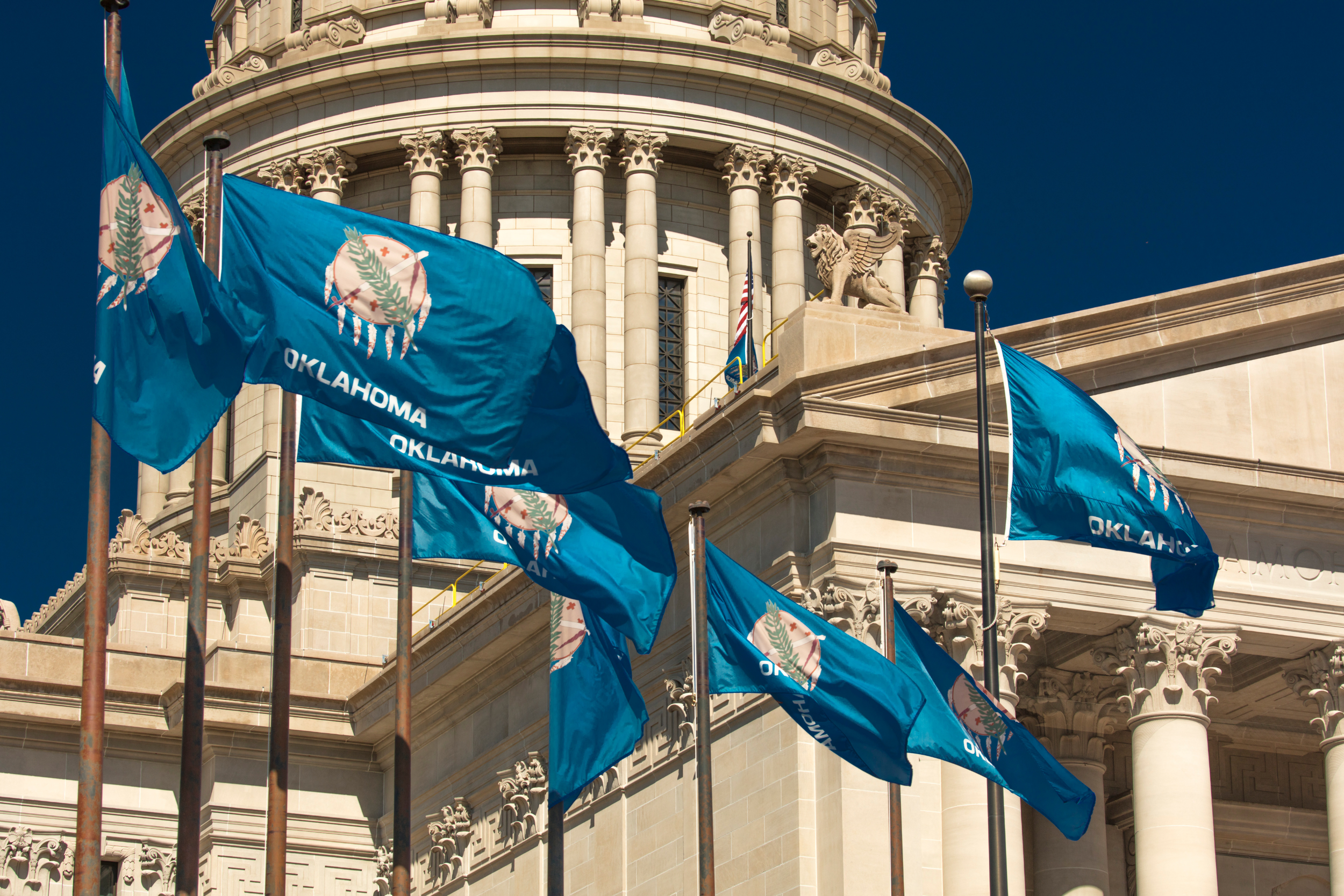 Flags with Oklahoma state emblem flying outside the statehouse building with columns and a lion sculpture on top, under a clear blue sky.