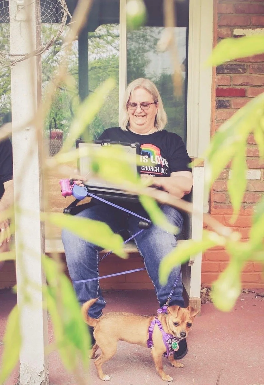 A woman with gray hair and glasses sitting outside on a porch, smiling while looking at a tablet, with a small tan dog on a purple leash in front of her. The porch has a brick wall and a large window, with green leaves in the foreground.