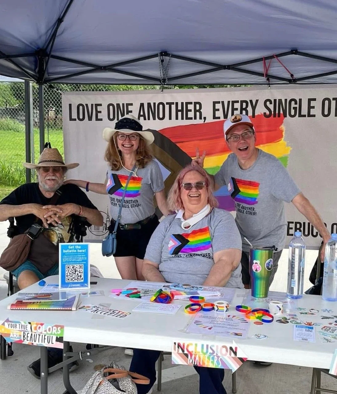 Four people smiling at an outdoor Pride event, wearing matching rainbow-themed shirts and hats, sitting and standing behind a table with Pride flags and informational flyers, under a tent with a banner that reads "Love one another, every single other".