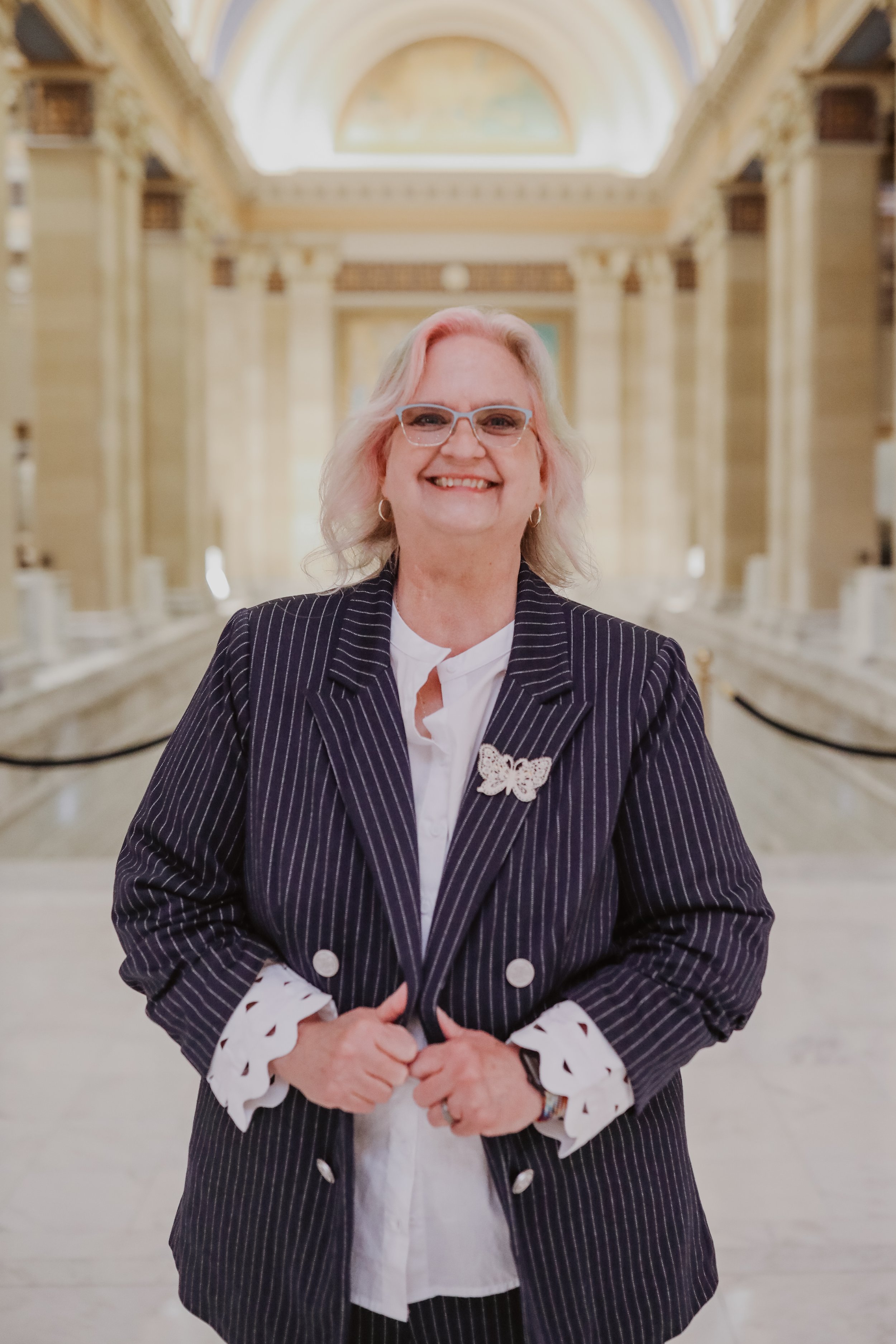 A woman,Vicki, wearing a navy pinstripe blazer with butterfly brooch, and white shirt smiling wildly inside the Oklahoma capital