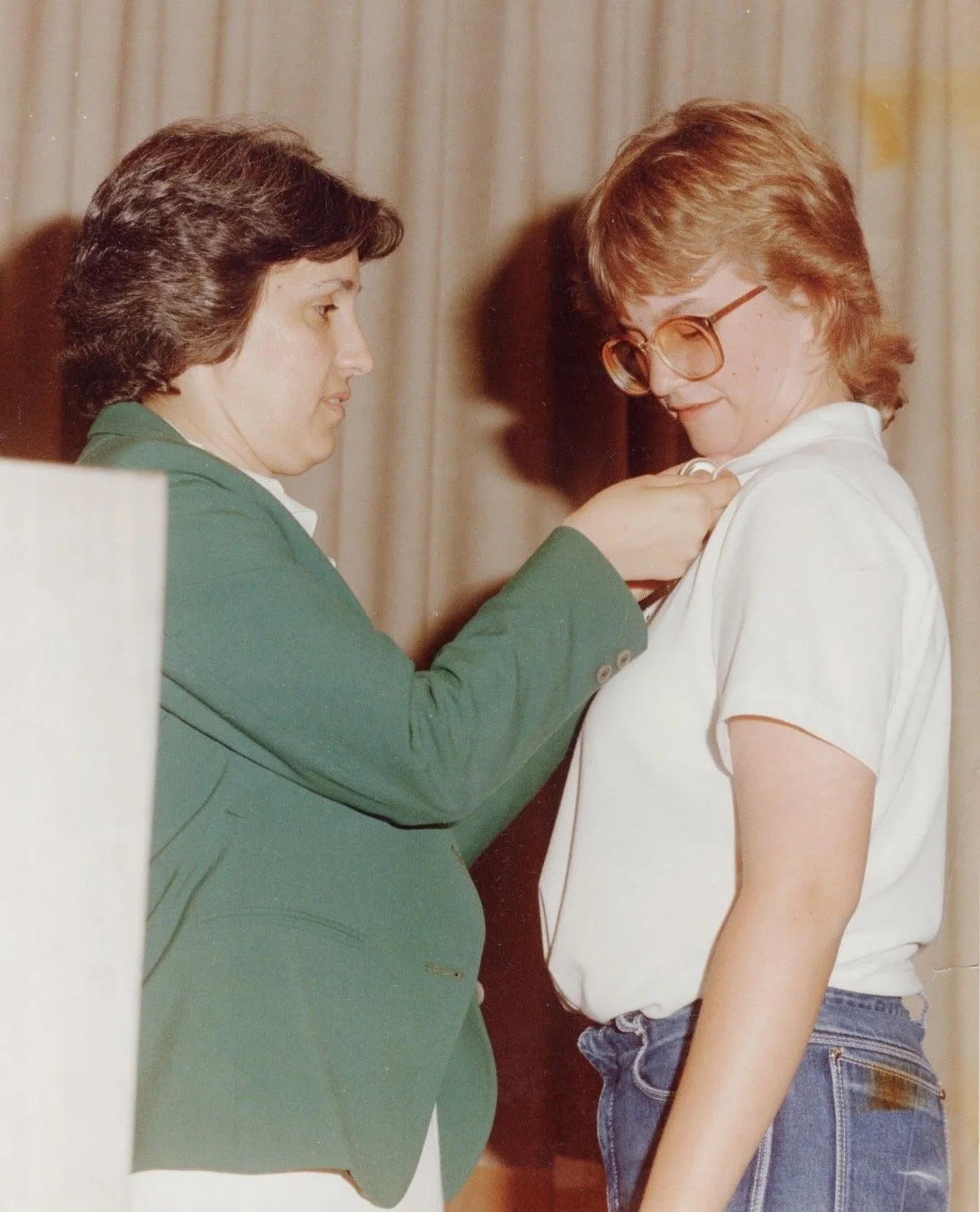 A woman in a green blazer pins a white shirt onto a young woman with glasses and jeans, as part of a religious or ceremonial event.