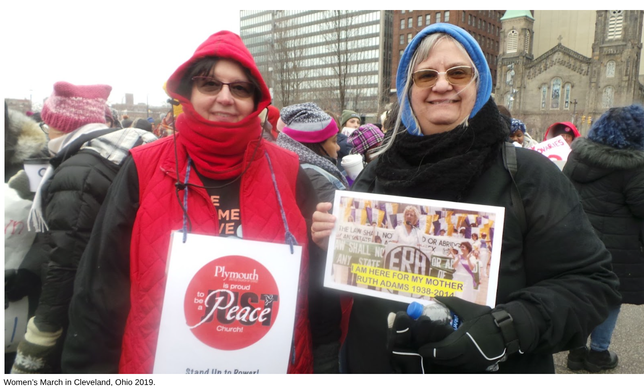 Two women standing together at a women's march in Cleveland, Ohio, 2019, holding signs and wearing warm clothing and glasses, with a crowd and city buildings in the background.