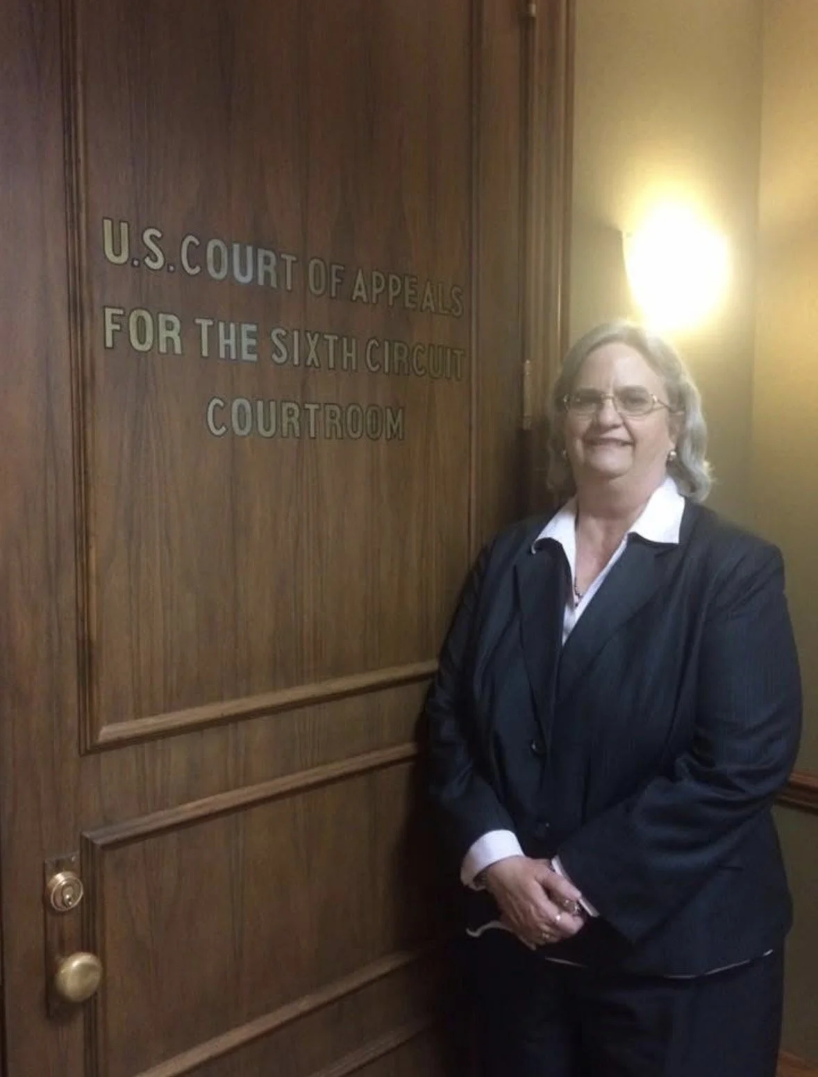 A woman in a dark suit standing next to a wooden door with the sign 'U.S. Court of Appeals for the Sixth Circuit Courtroom'.