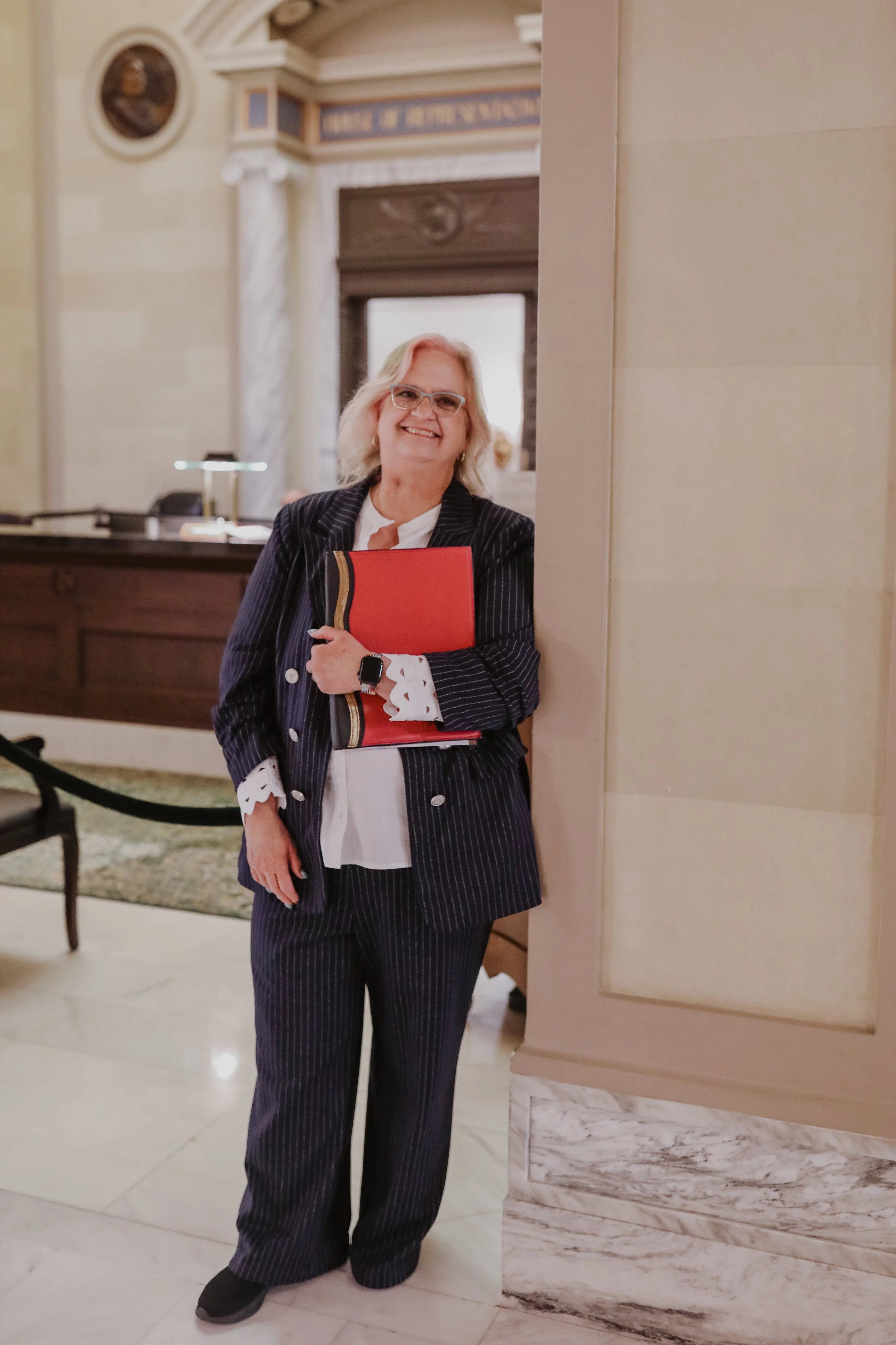 A woman in a pinstripe suit holding a red folder, standing in a government building lobby, smiling.