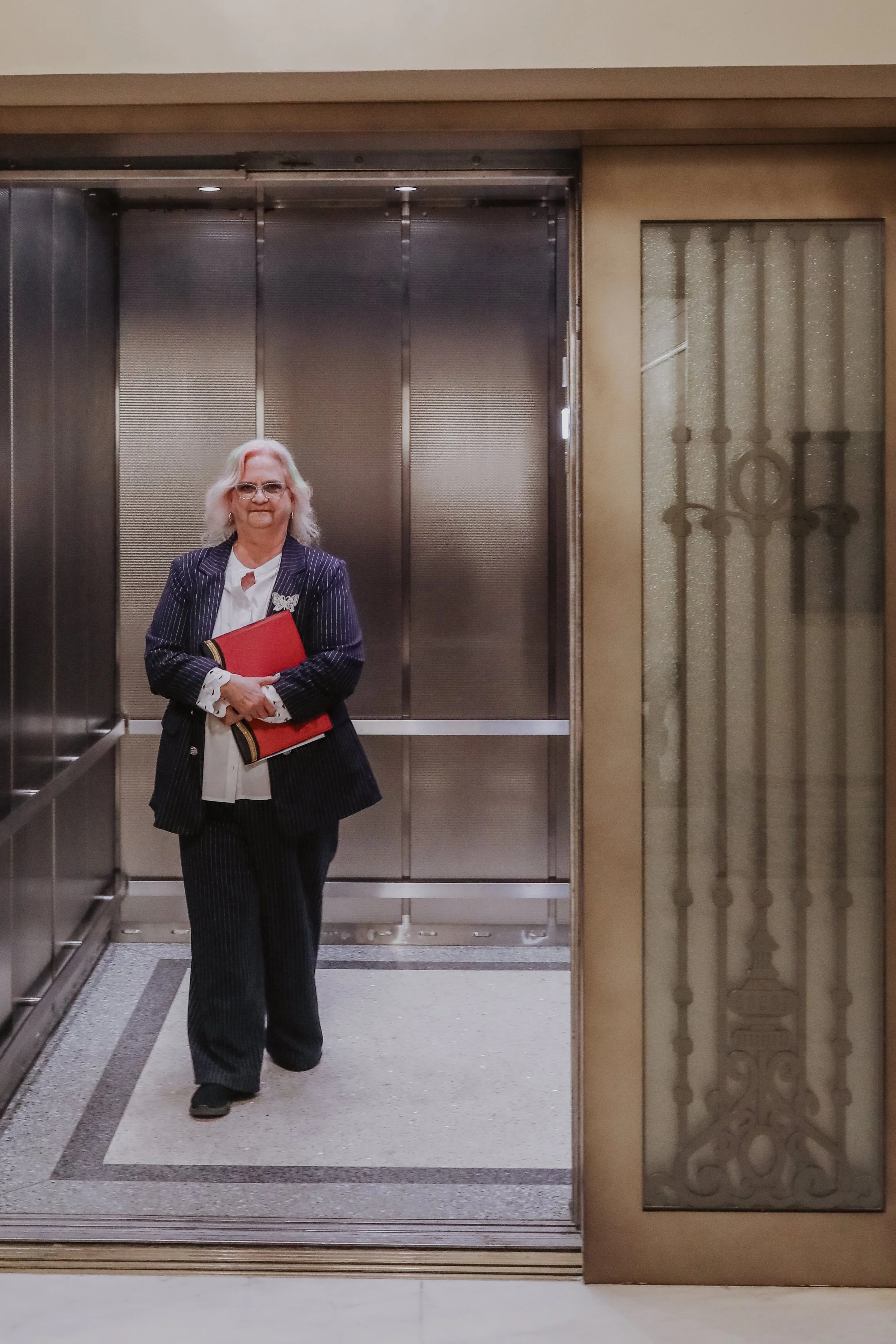 Vicki in a pinstripe suit holding a red folder is standing inside an elevator with stainless steel doors, in front of a decorative grill on the right.
