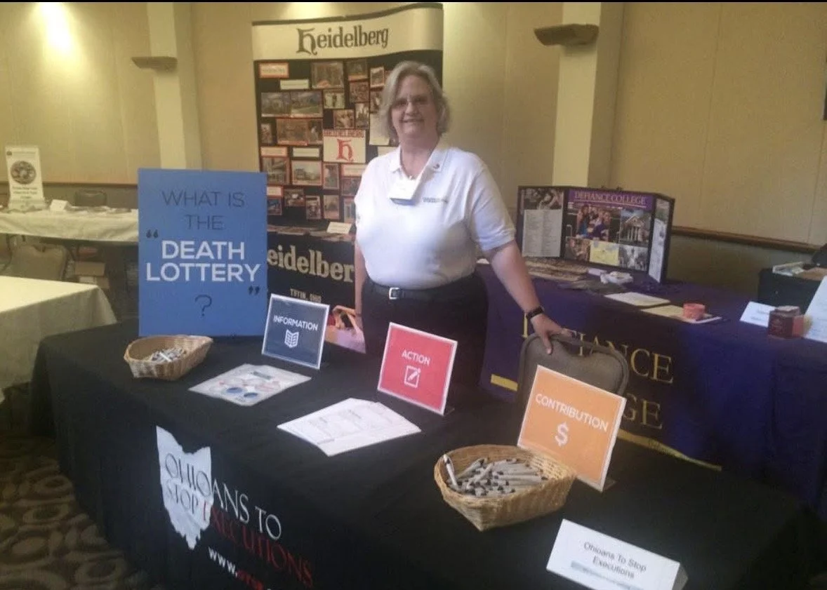Woman standing behind a table at an informational booth about the death lottery, with signs labeled 'Information,' 'Action,' and 'Contribution,' and a backdrop displaying 'Heidelberg.'