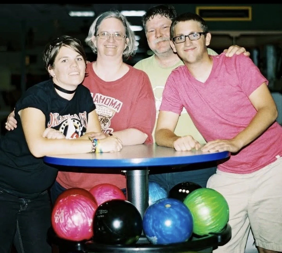 Four people in a bowling alley, standing around a table with colorful bowling balls underneath. They are smiling and appear to be enjoying a fun outing.