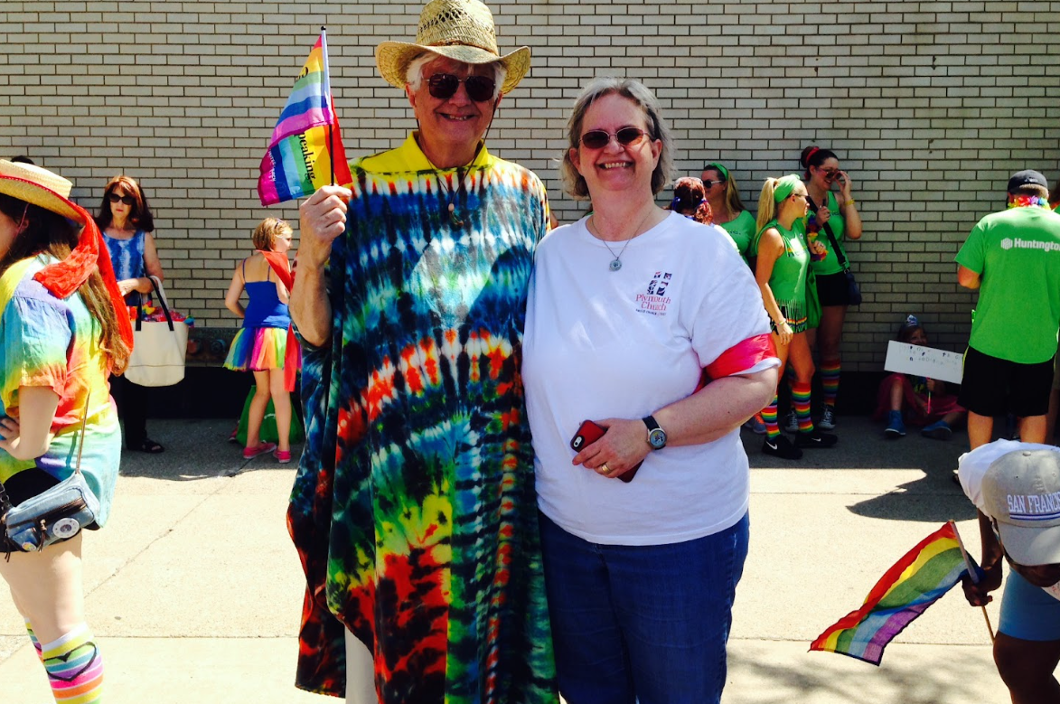 Two smiling people standing together at a pride parade with rainbow decorations and flags, wearing colorful and casual clothing.