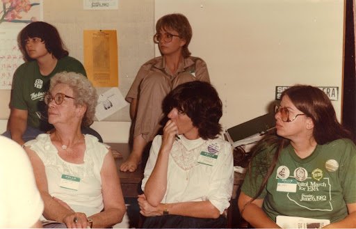 Four women and two men in a room, some wearing shirts with messages, appearing to be at a meeting or event.