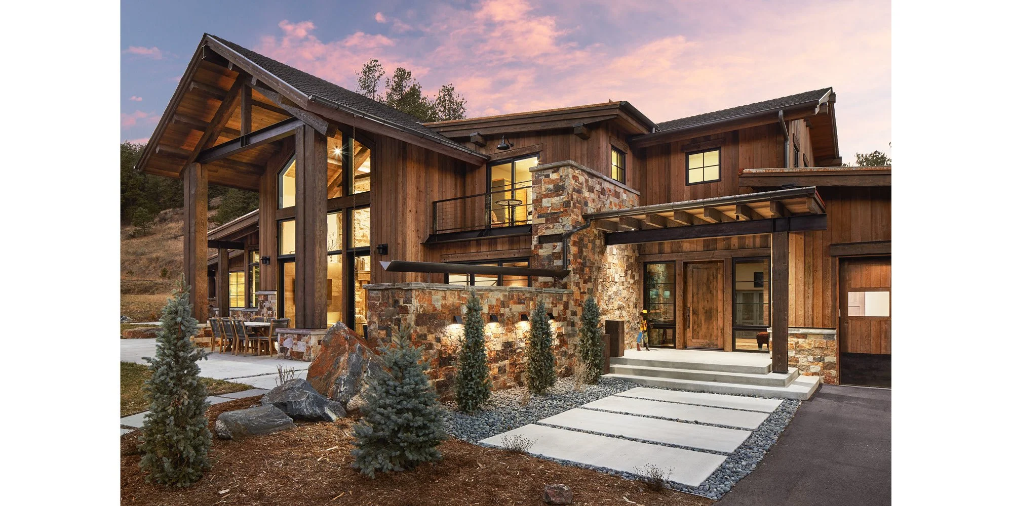 A modern house with wooden and stone exterior walls, large glass windows, and a balcony, set against a sunset sky.