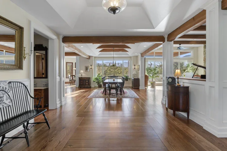 Spacious open-concept living room with hardwood floors, white walls, wooden beams, and large windows with a view outside.