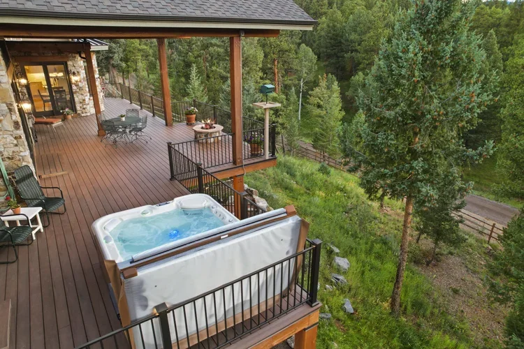 A wooden deck with a hot tub, outdoor furniture, and lush green trees and forest in the background.