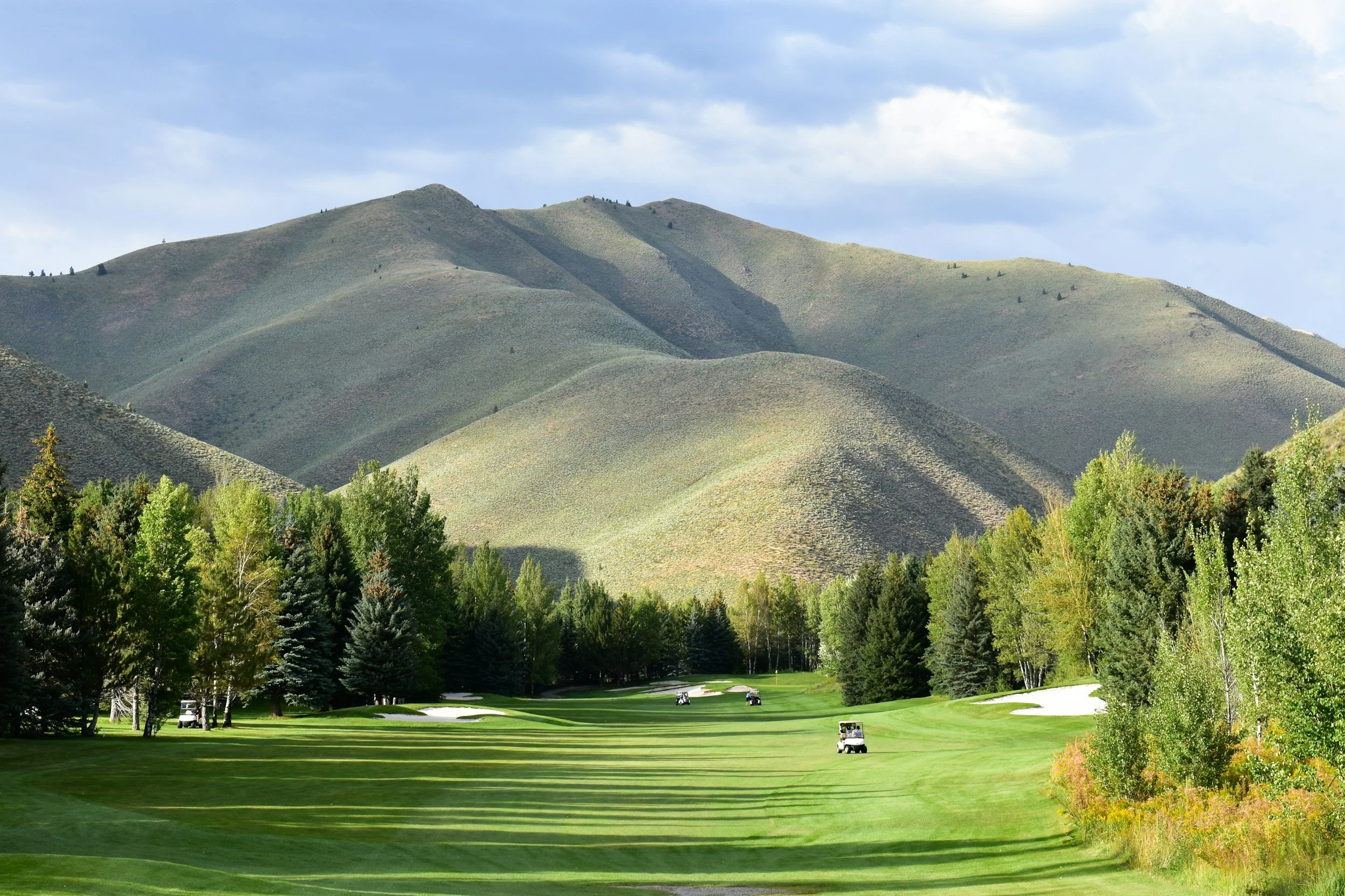 A scenic view of a golf course surrounded by lush trees with a mountain range in the background under a partly cloudy sky.