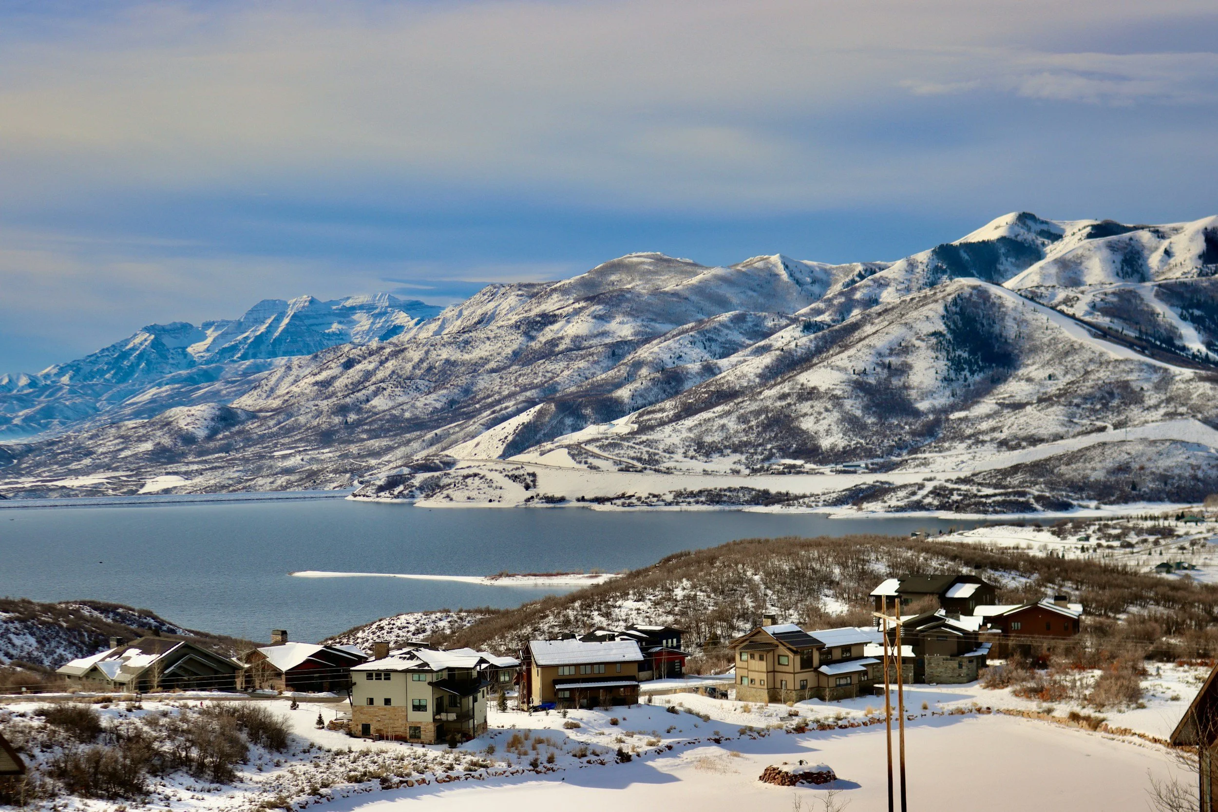 Snow-covered mountains with a lake in the foreground, and a small residential area with houses at the base of the mountains, under a partly cloudy sky.