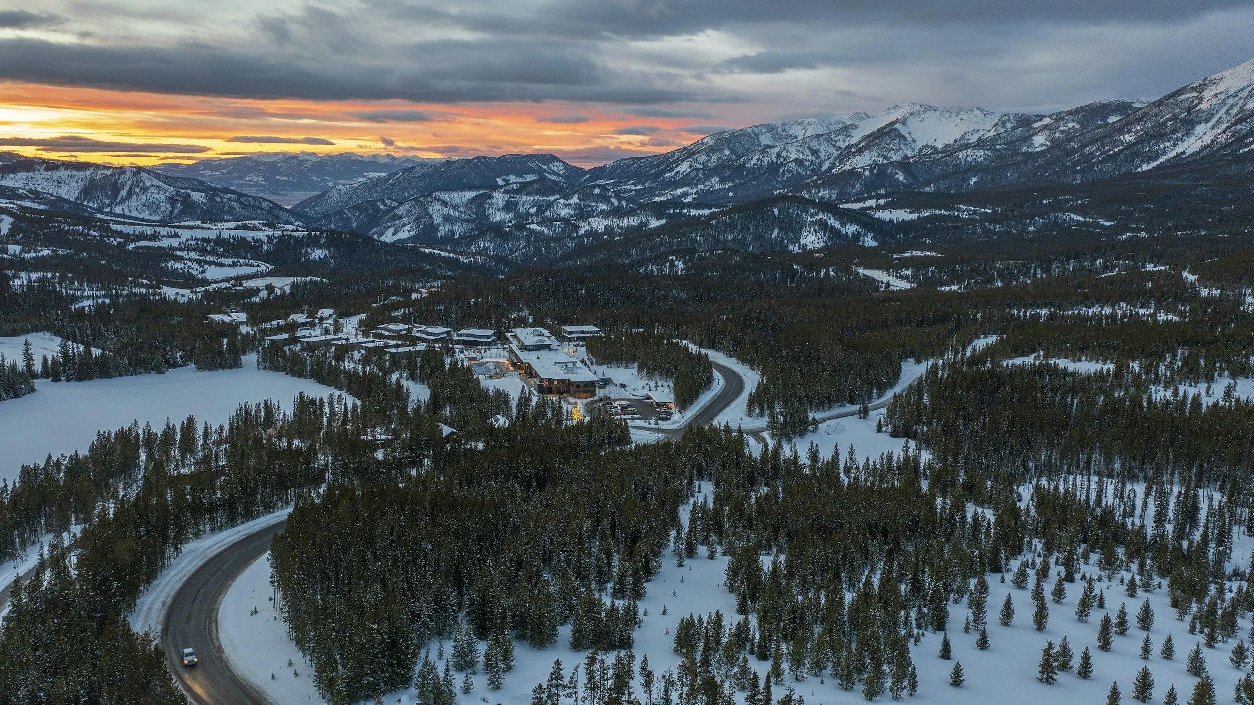 Snow-covered mountains and forest at dusk with a small, illuminated town and winding road in the foreground.