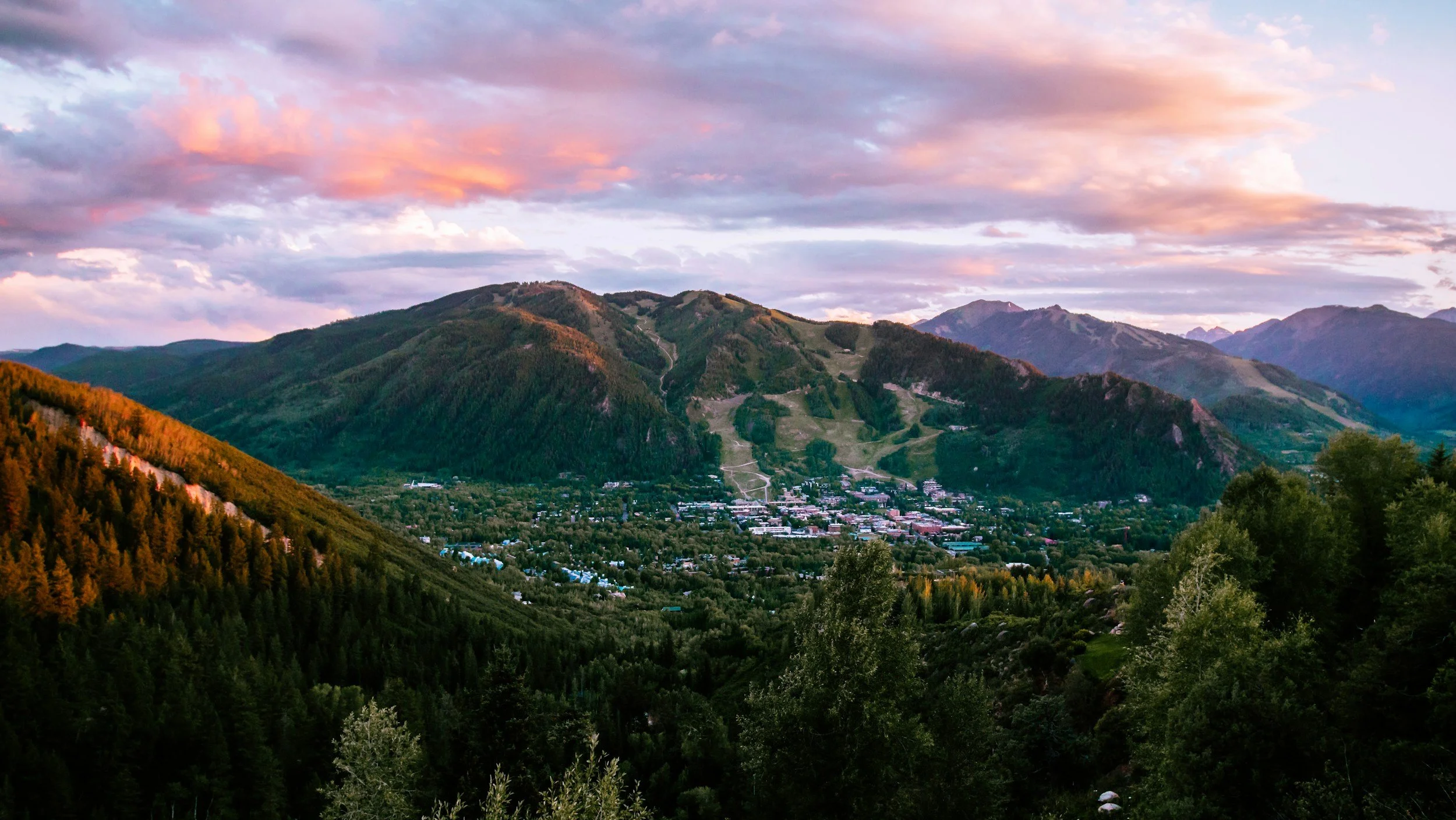 Scenic view of green mountains at sunset with pink and purple clouds in the sky and a small town nestled in the valley.