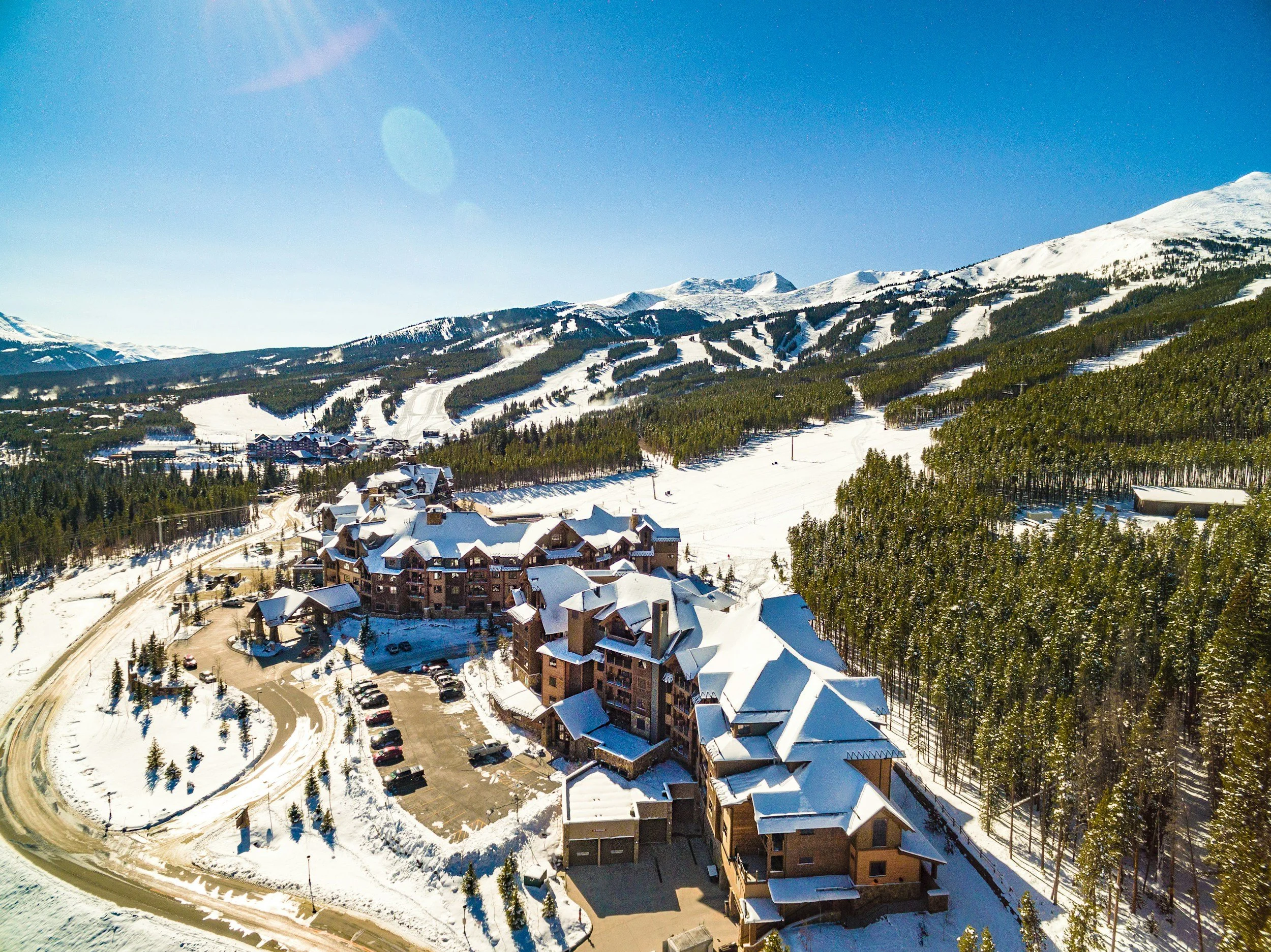 An aerial view of a snowy mountainous landscape with a small town or resort at the base, surrounded by green trees and ski slopes under a clear blue sky.
