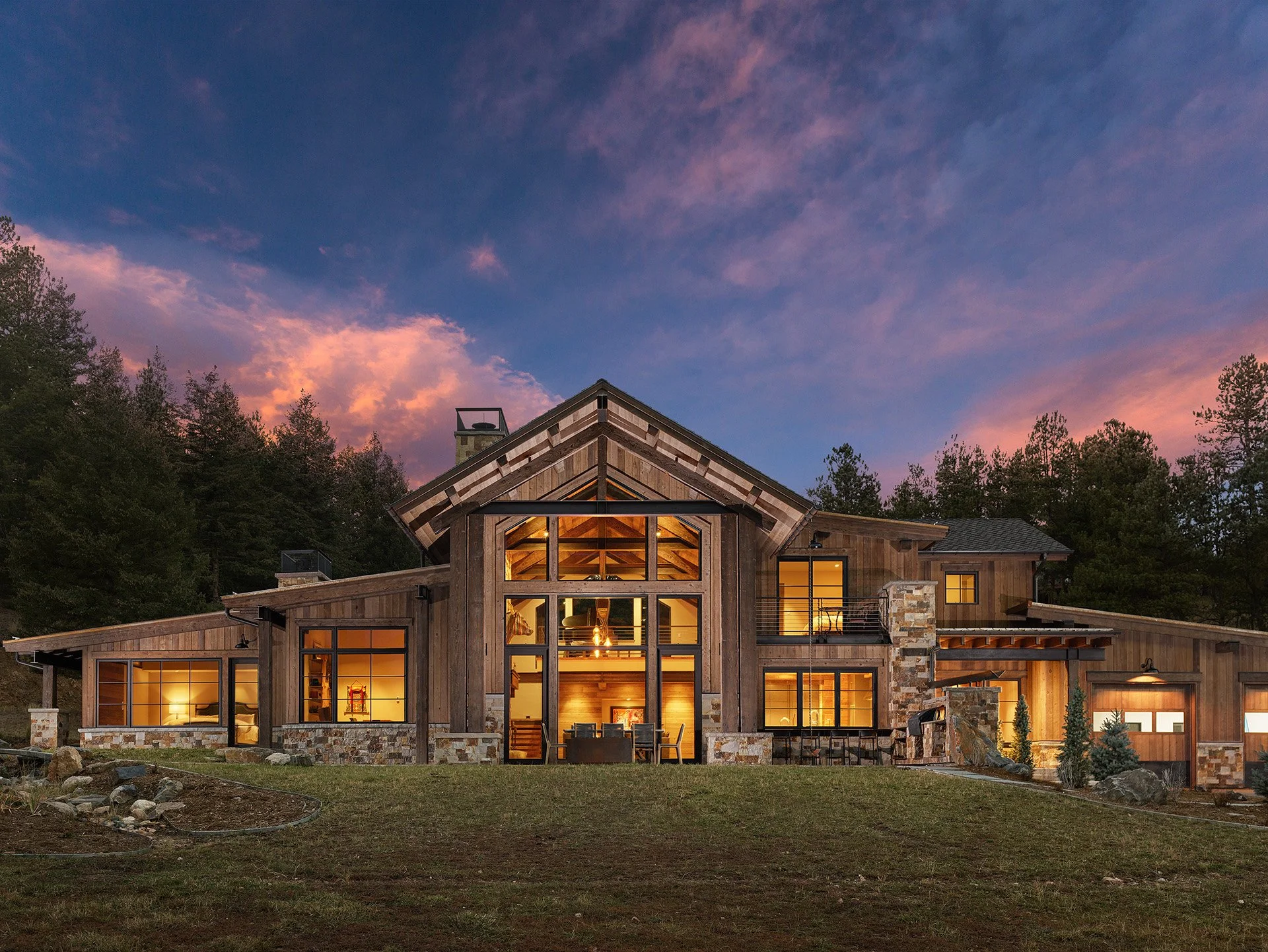 A modern house with large glass windows illuminated from inside, surrounded by trees at sunset or dusk, with a colorful sky featuring pink and purple clouds.