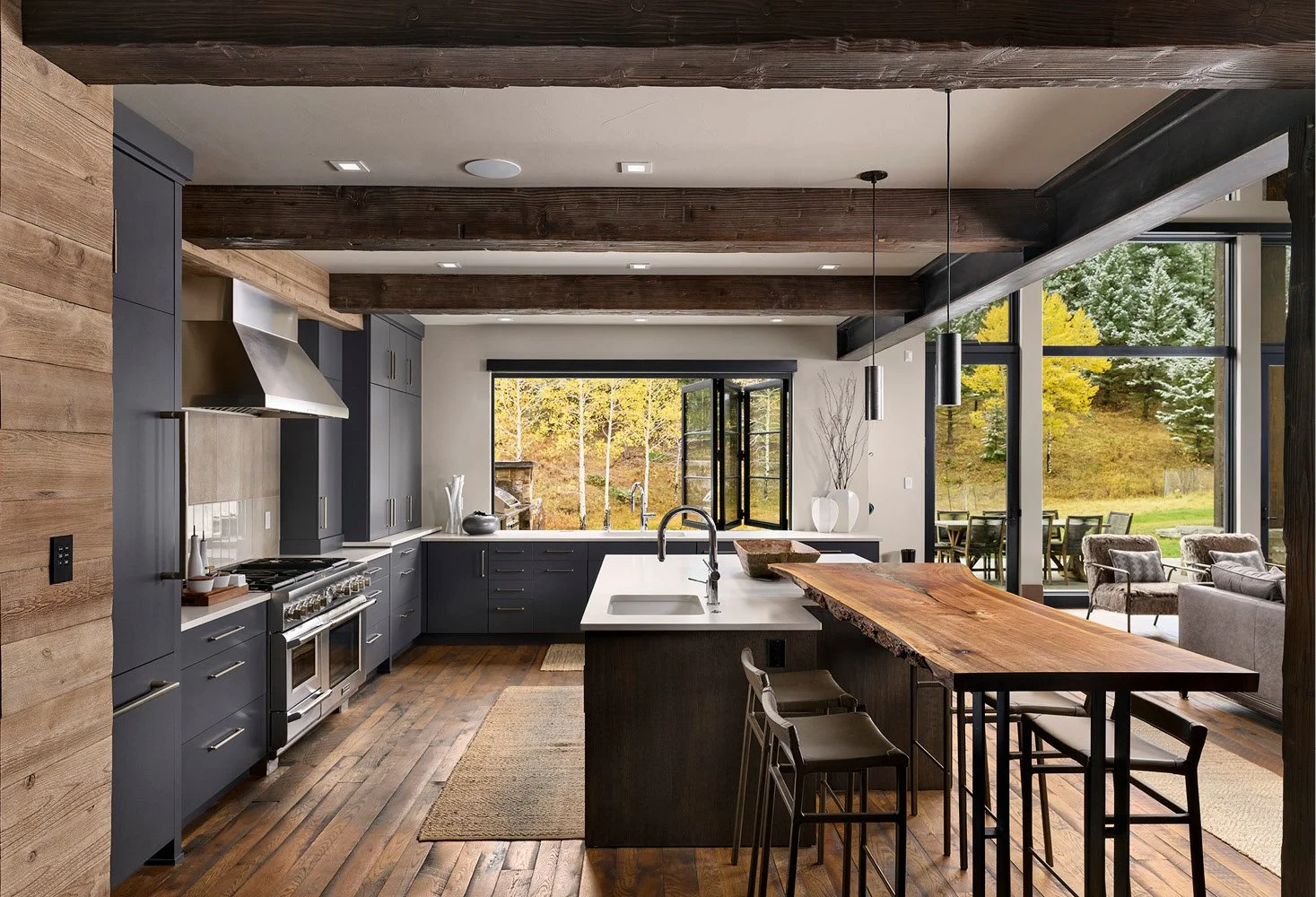 Modern kitchen with dark blue cabinets, wooden ceiling beams, a large island with a wood top, and large windows overlooking a green outdoor space.