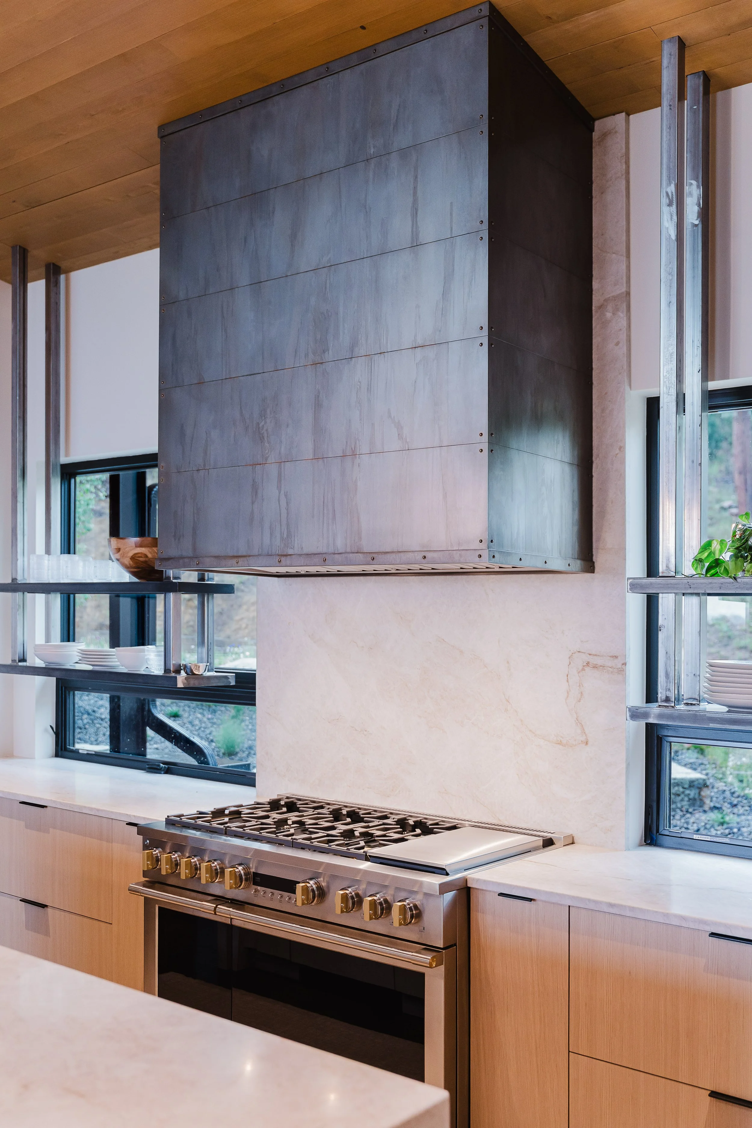 Modern kitchen with a stainless steel stove, wooden cabinets, and a stainless steel vent hood above the stove. There are large windows with black frames, and the countertop is made of light-colored stone.