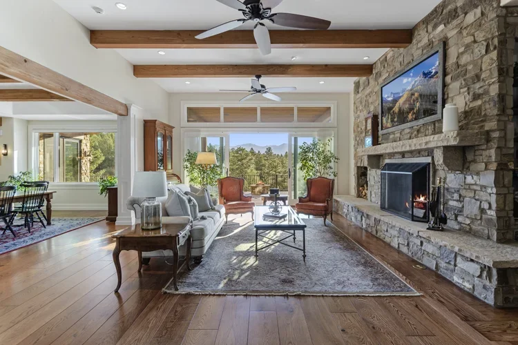 Living room with large stone fireplace, flat-screen TV, wooden beams on ceiling, hardwood floors, and large window with views of trees and mountains.