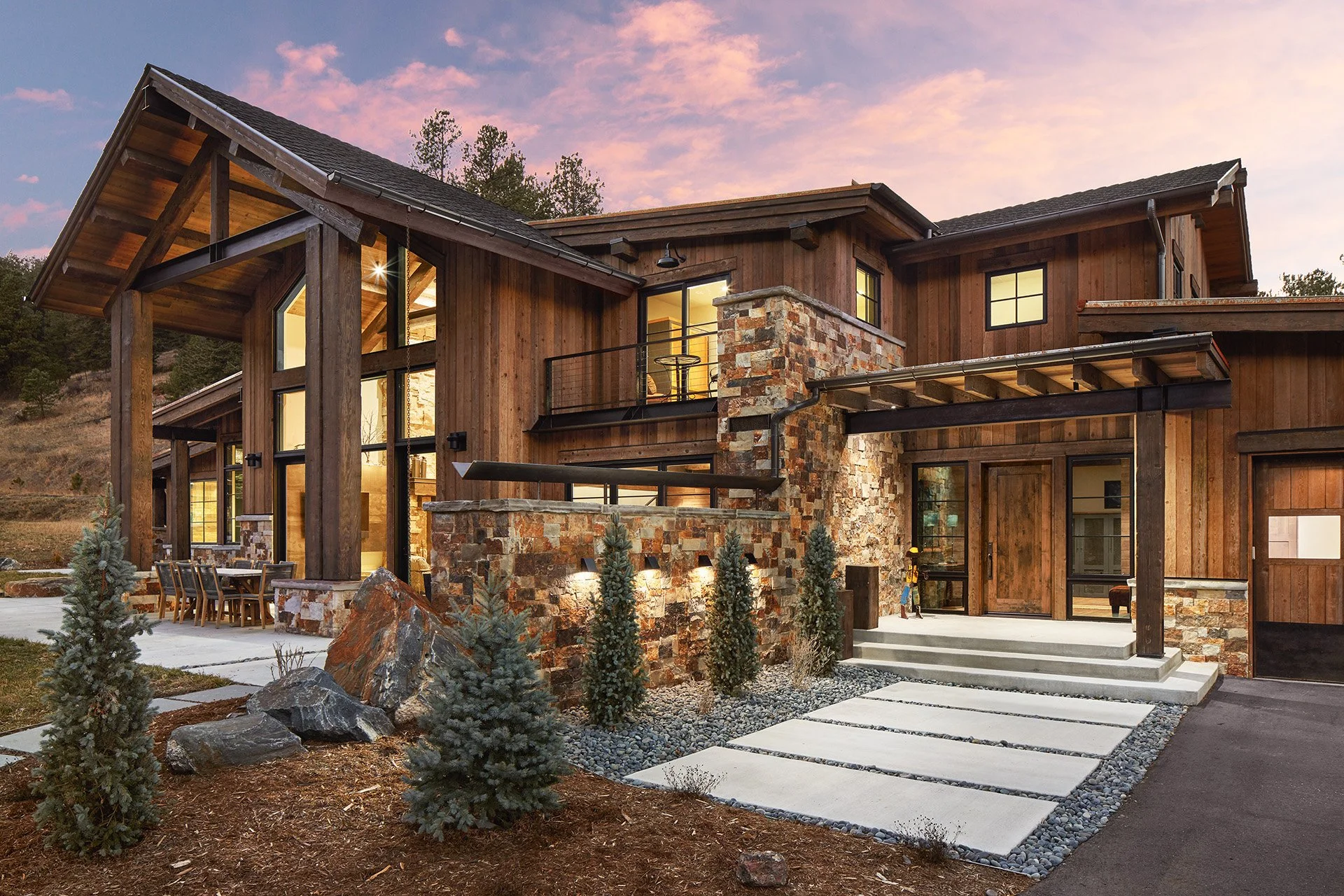 Modern house with wooden siding and stone accents, large windows, outdoor seating area, landscaped yard with small trees and rocks, set against a sunset sky.