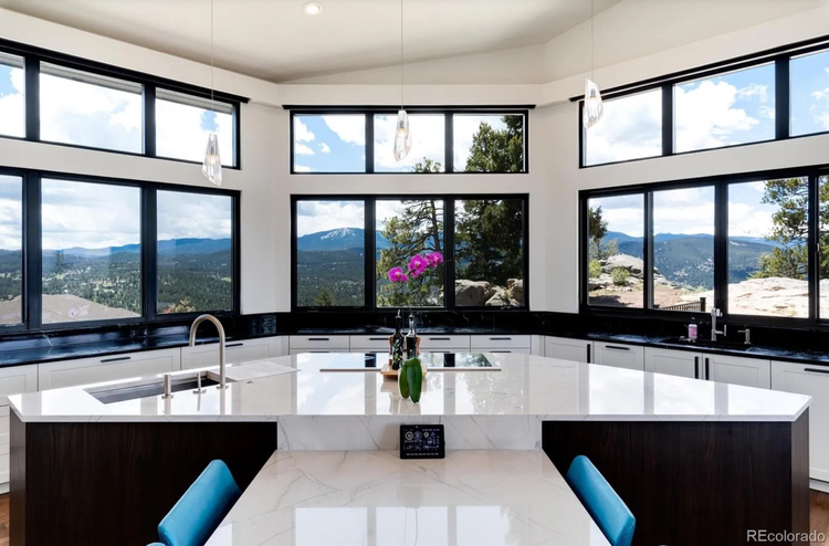 Modern kitchen with black countertops, white cabinets, and large panoramic windows showing mountains and trees outside.