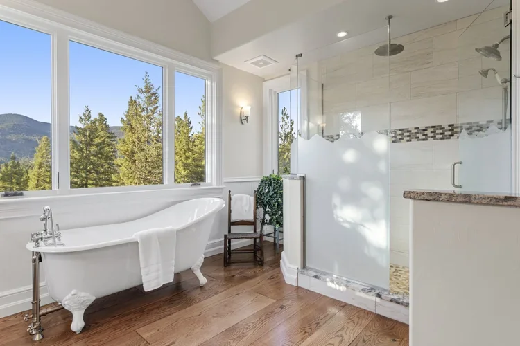 Bright bathroom with a clawfoot bathtub, large windows with a view of trees and mountains, and a glass-enclosed shower with modern fixtures.