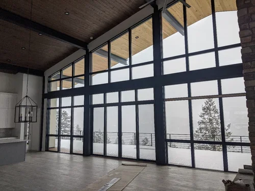 Large windows revealing an outdoor view of trees and snow, inside a modern room with hardwood ceiling and floor, a hanging lantern, and a brick wall.