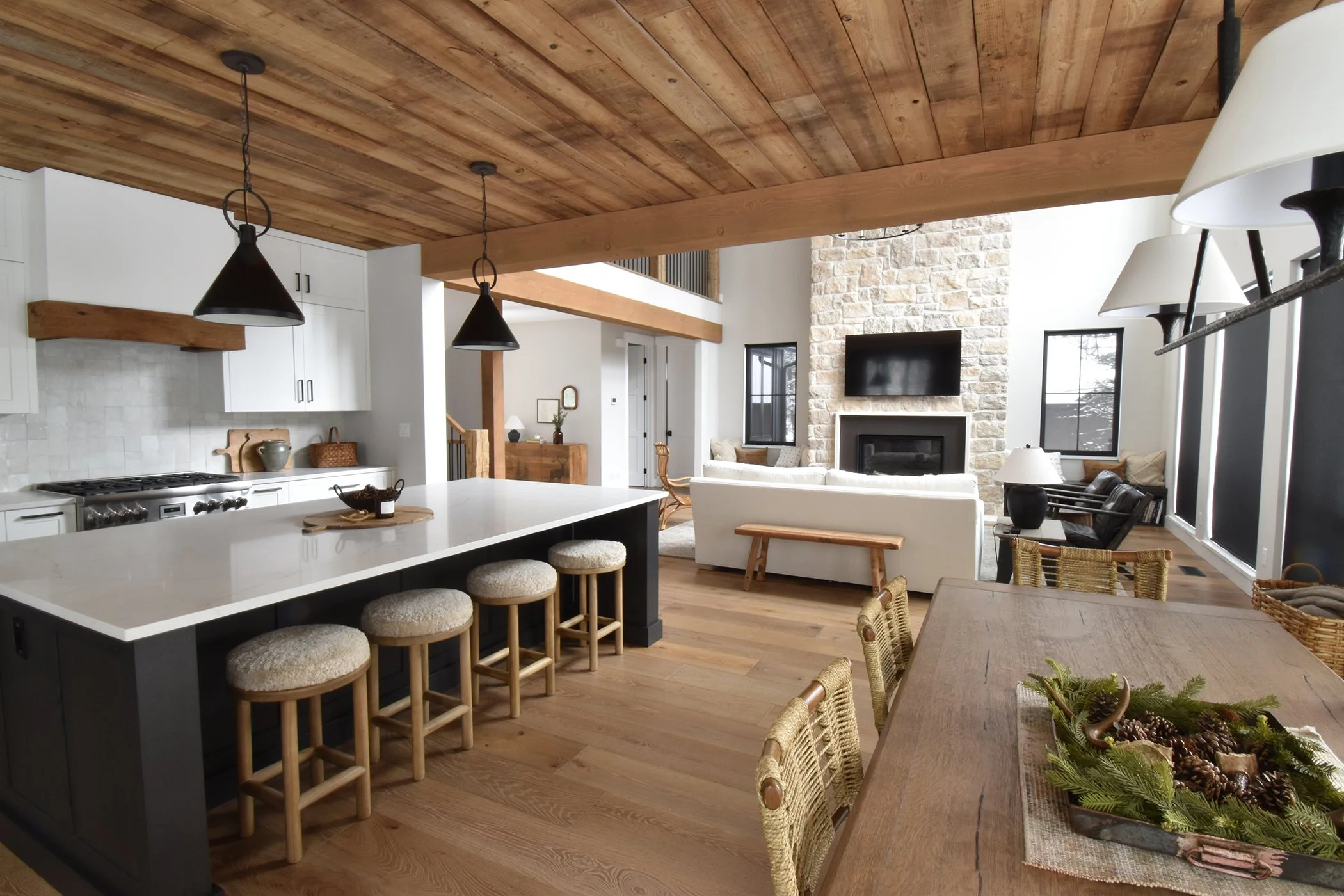 Open-concept living area with kitchen, dining, and sitting spaces featuring wooden ceiling, stone fireplace, flat-screen TV, lounge chairs, and large windows.