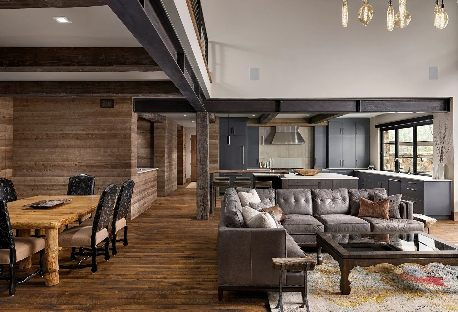 Open-concept living and dining area with wood floors, a brick wall, a gray kitchen with black cabinetry, a large window, and a gray sofa with pillows.